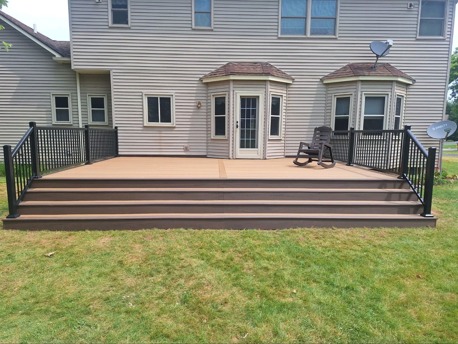 Backyard deck with brown steps, black railing, and a rocking chair. House with satellite dishes in the background.