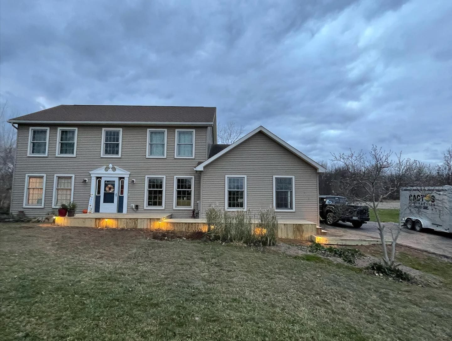 Two-story house with light-colored siding, a front porch with lit-up lights, and a cloudy sky.