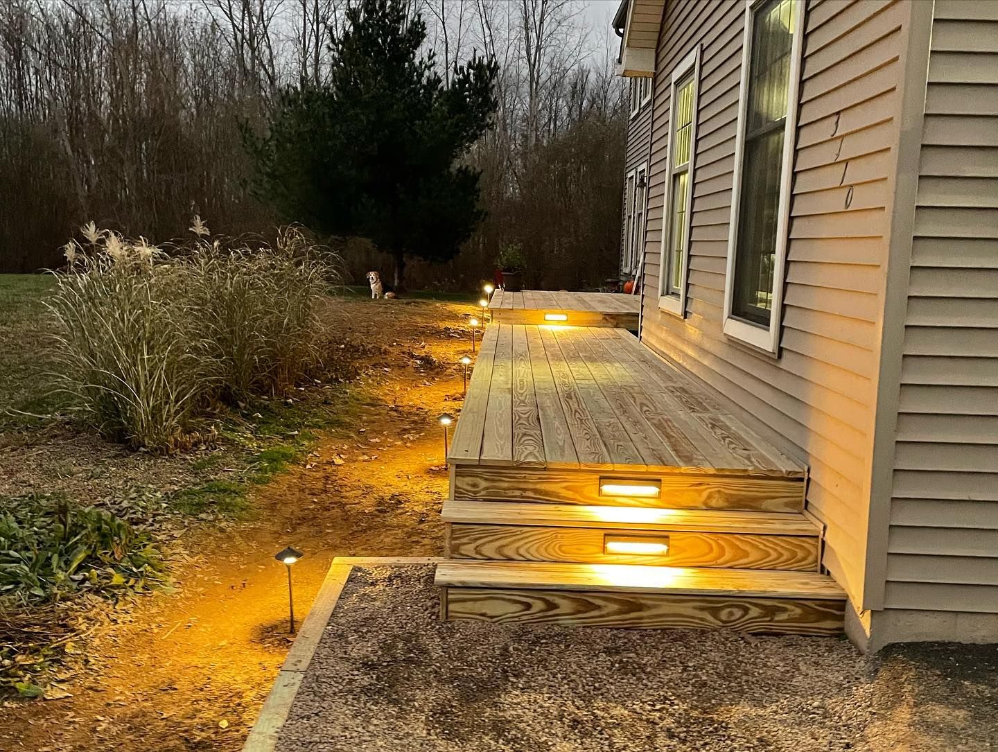 Lit wooden deck and pathway alongside a tan house at dusk.  Warm lights illuminate steps and walkway.