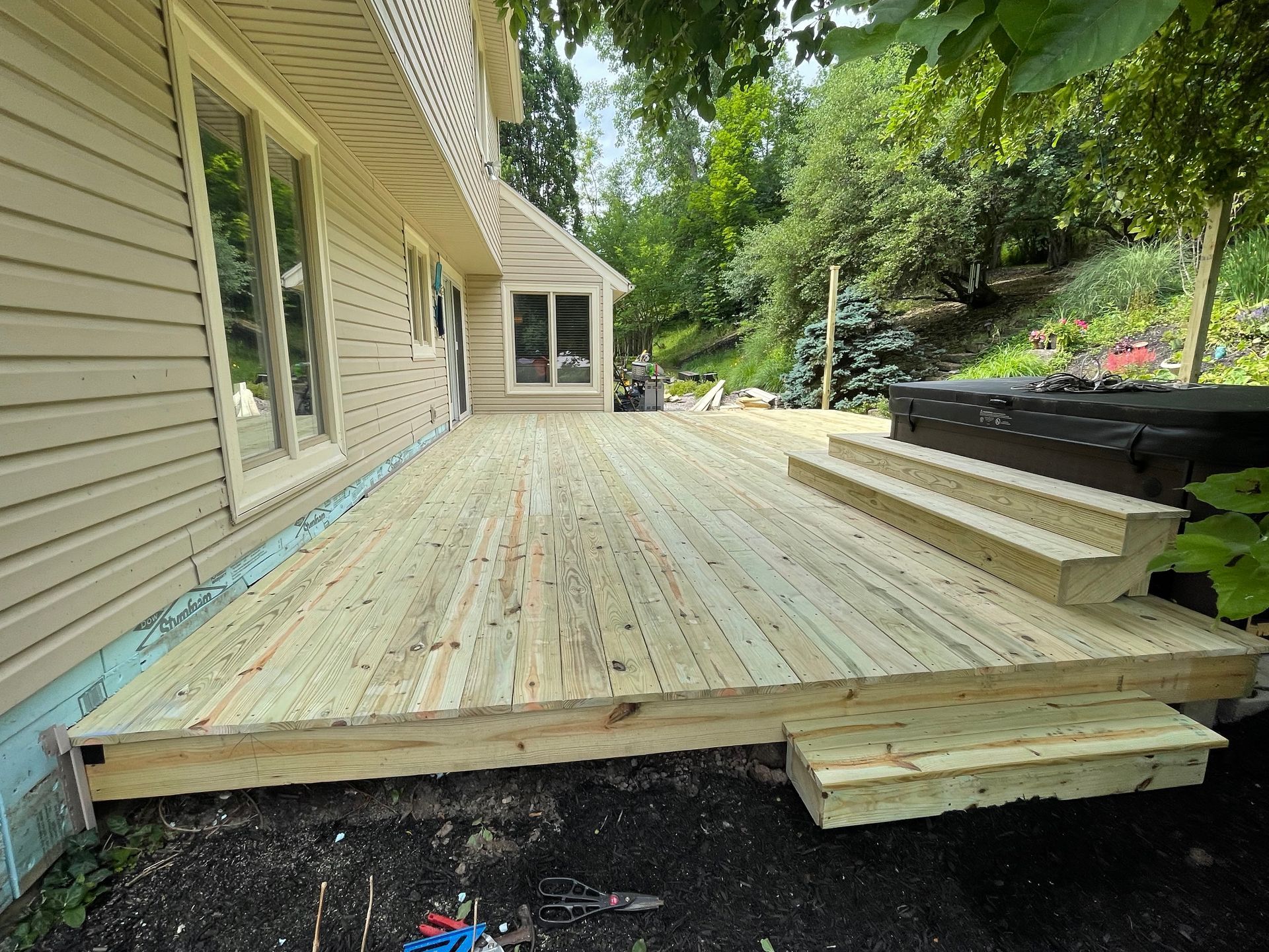 Newly built wooden deck attached to a beige house, with steps leading to a hot tub in a backyard setting.