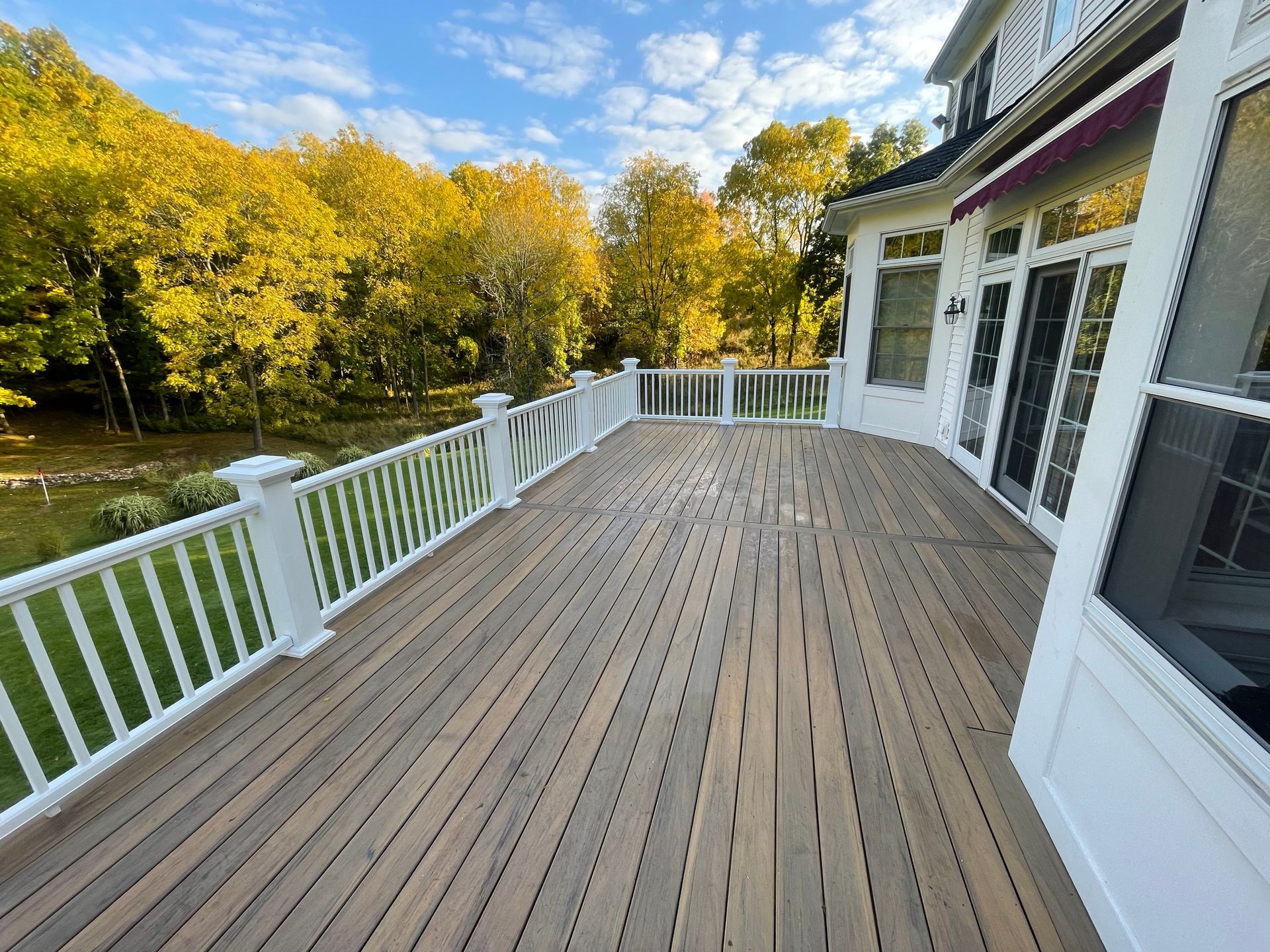 Wooden deck with white railing next to a house with large windows, autumn trees in background.