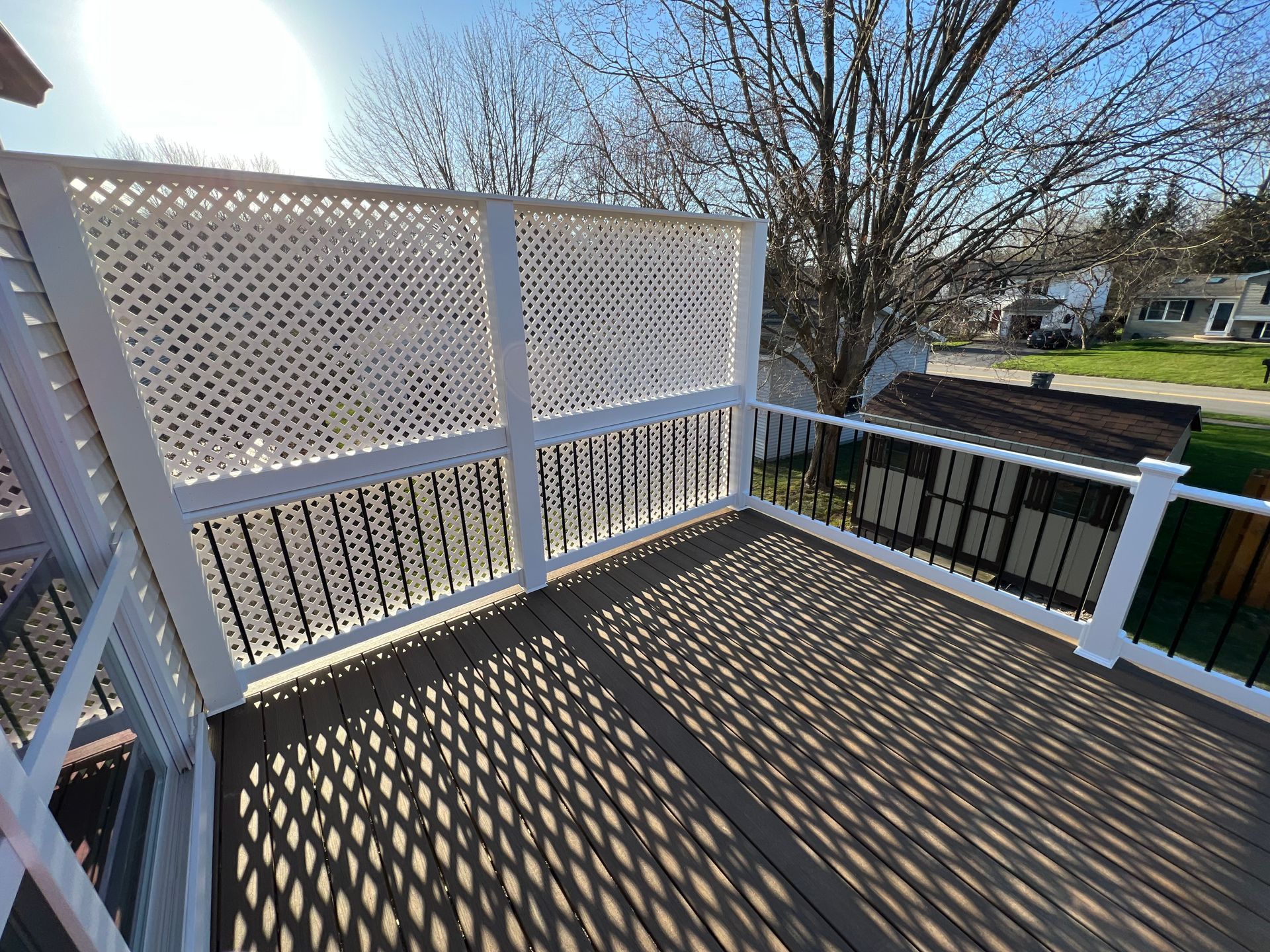 Deck with lattice screen, casting shadows on the wooden planks; view of yard and houses.
