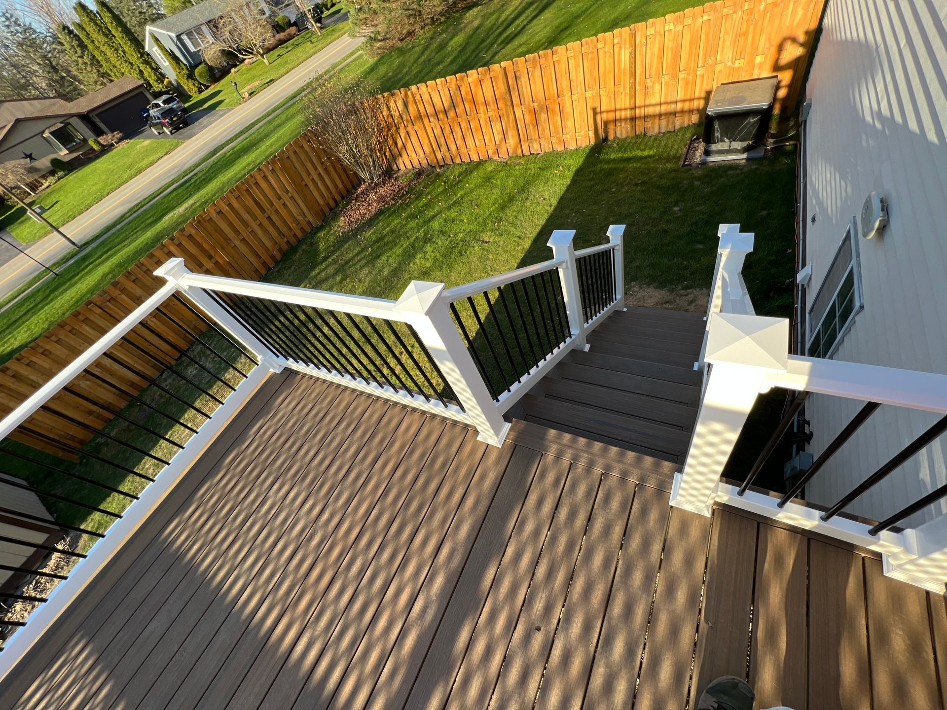 Wooden deck with stairs leading down to a grassy backyard, a brown fence, and houses.