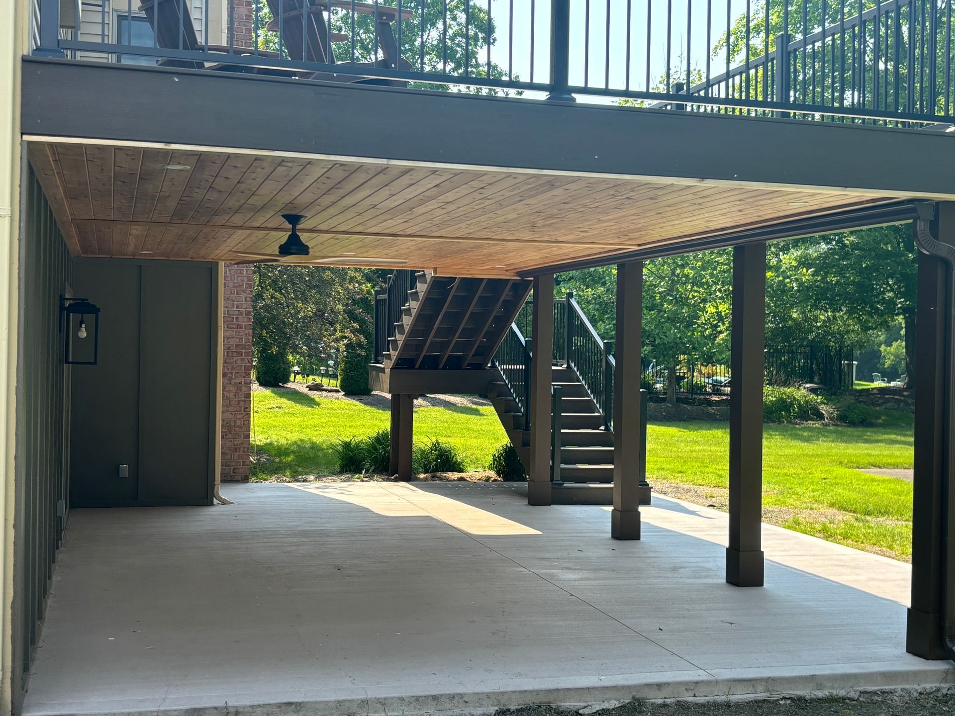 Concrete patio under a deck with stairs, a ceiling fan, and green lawn.