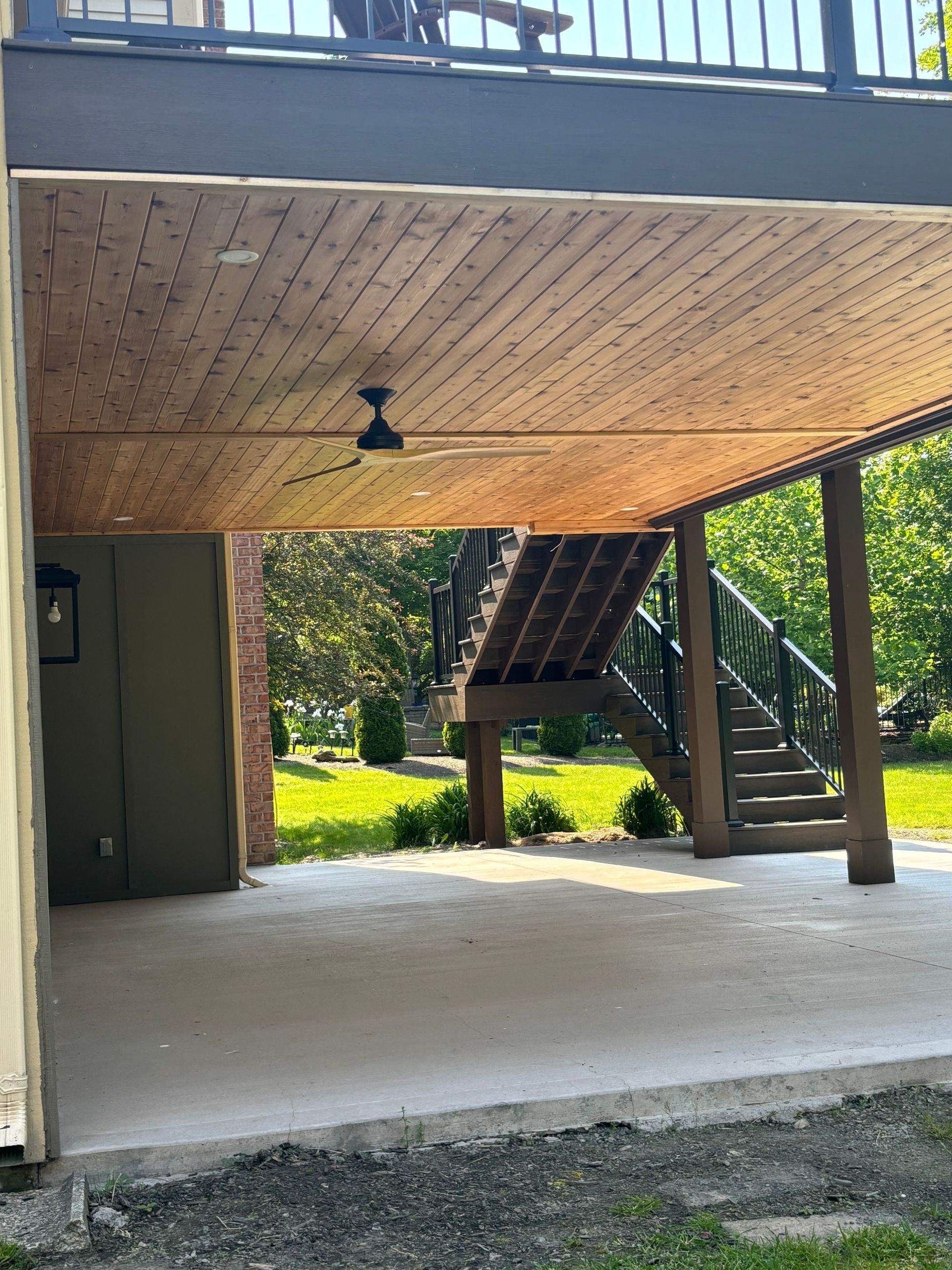 A concrete patio under a wooden deck with stairs, ceiling fan, and greenery.
