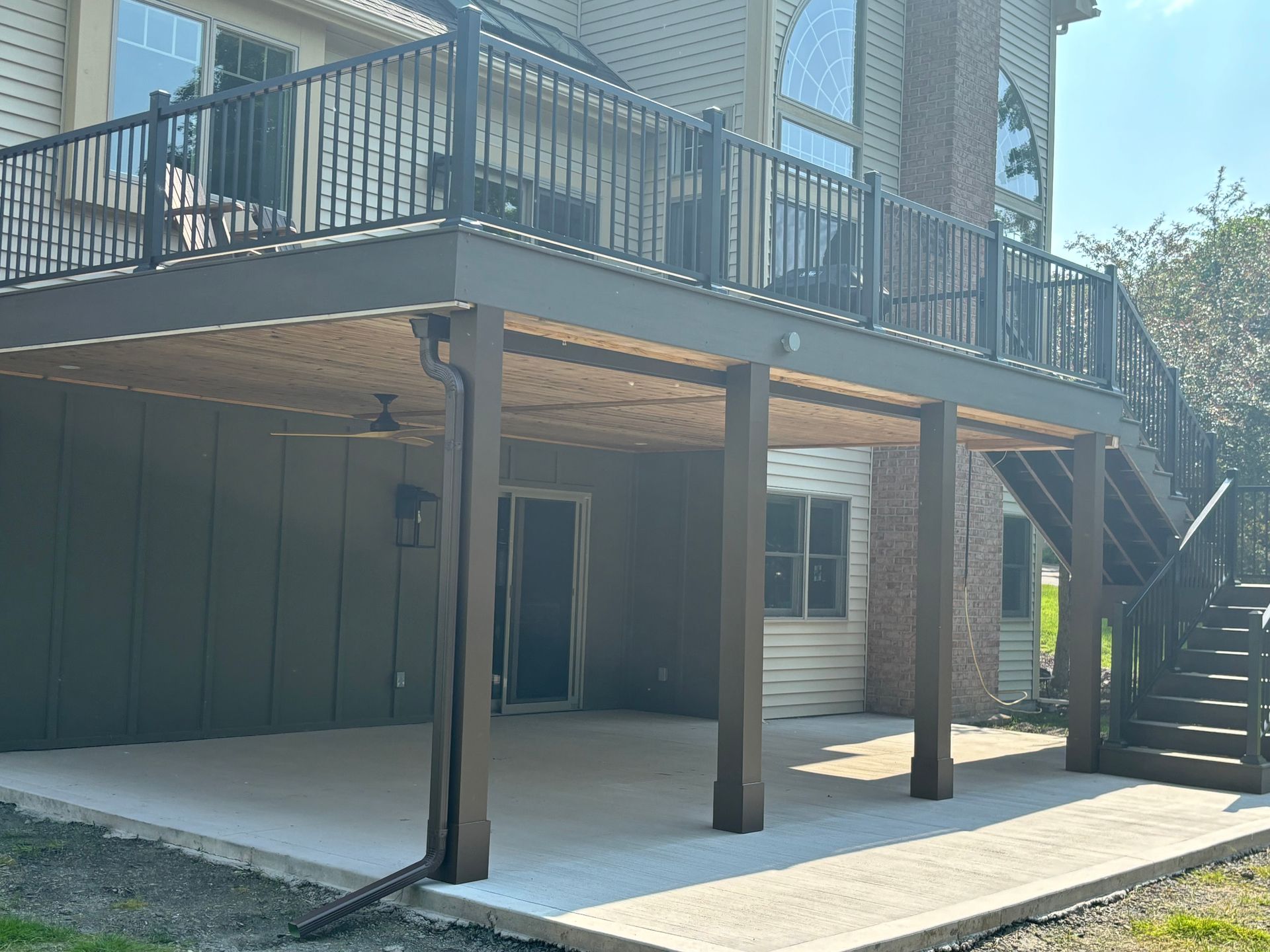 Two-story deck with stairs and concrete patio beneath, supported by brown posts, overlooking a yard.