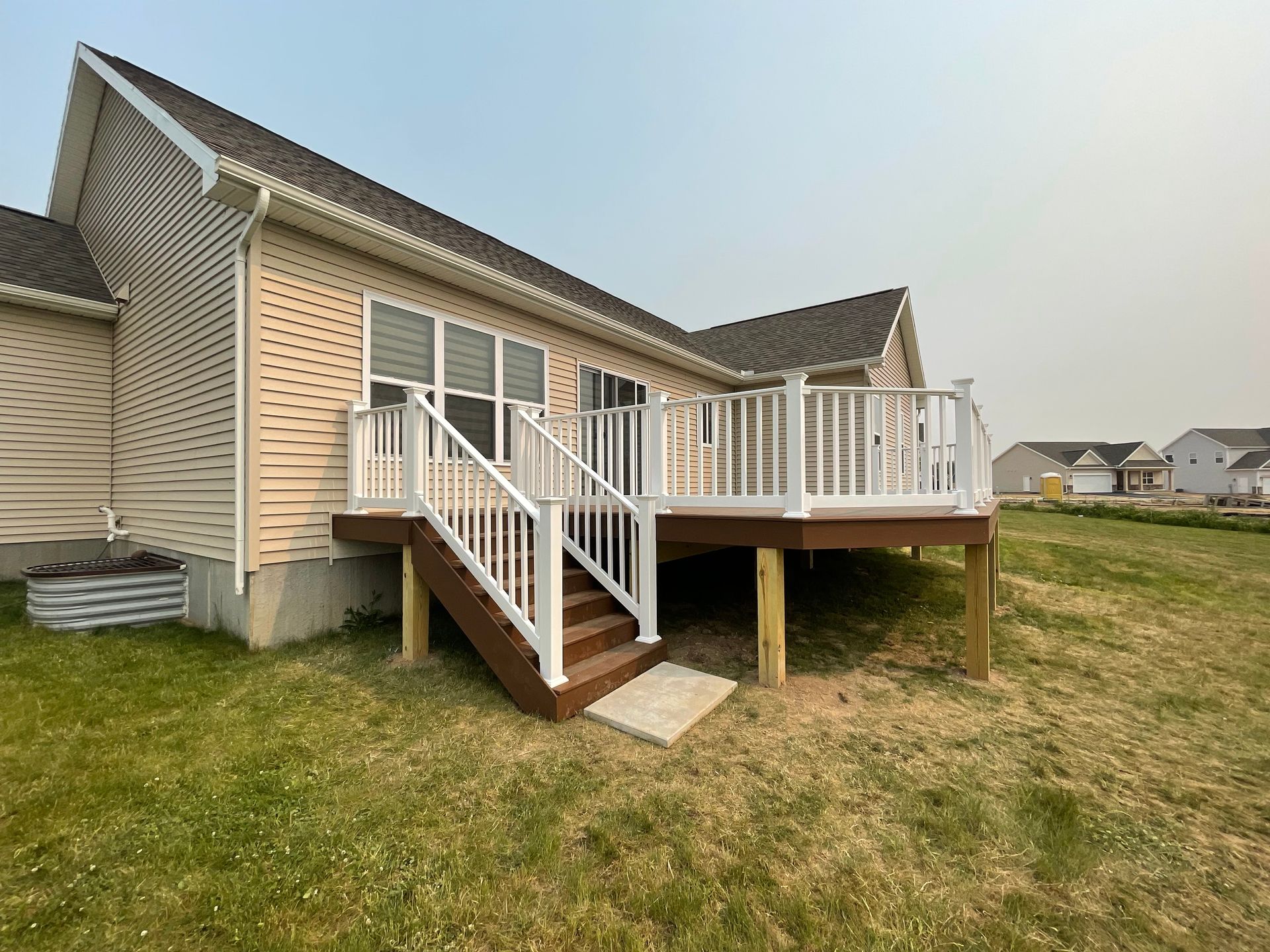 A wooden deck with white railing, attached to a tan house. Green grass surrounds the deck.