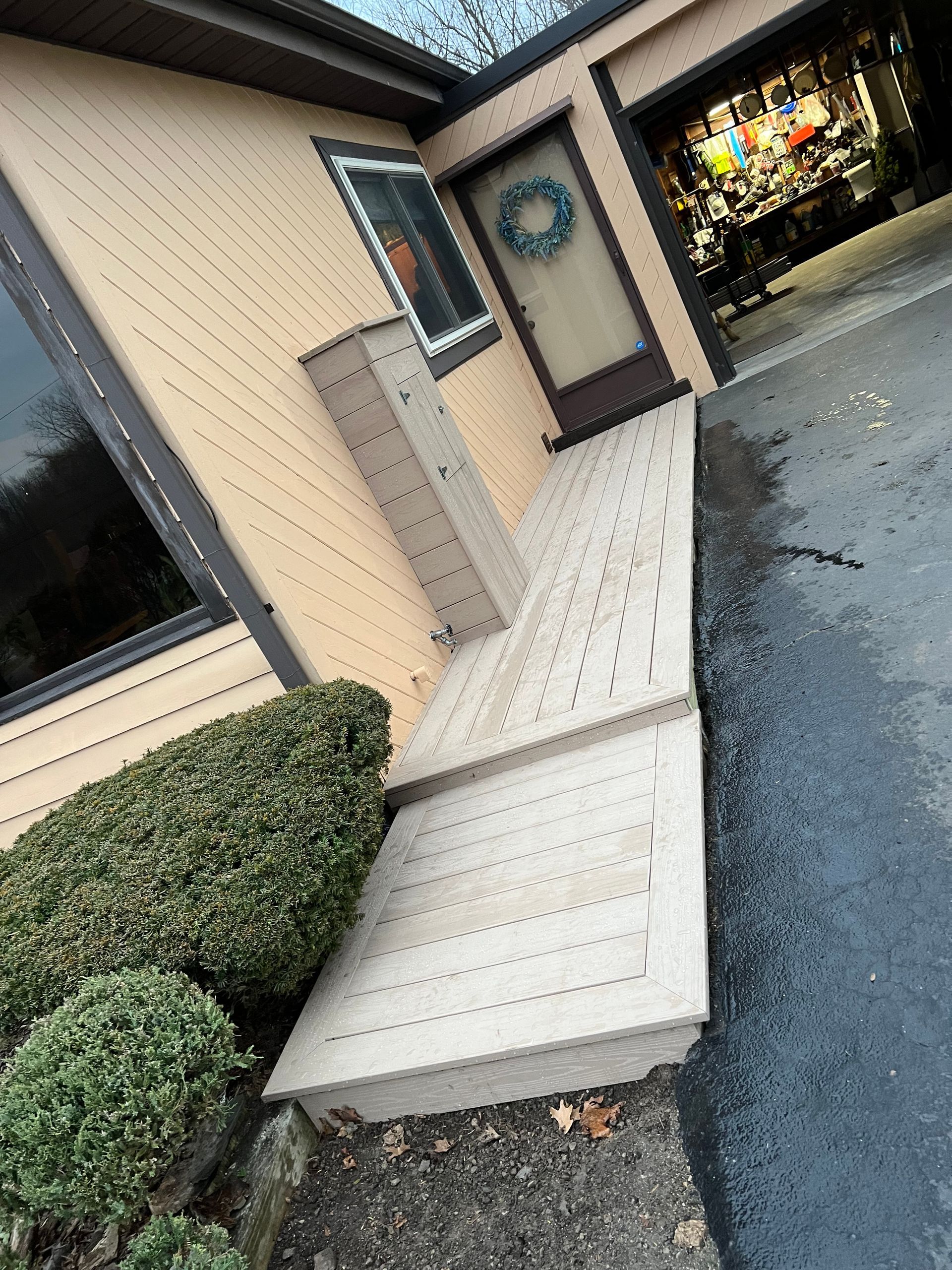 Wooden ramp leading to a doorway, alongside beige building with dark trim. Includes a small window and a garage.