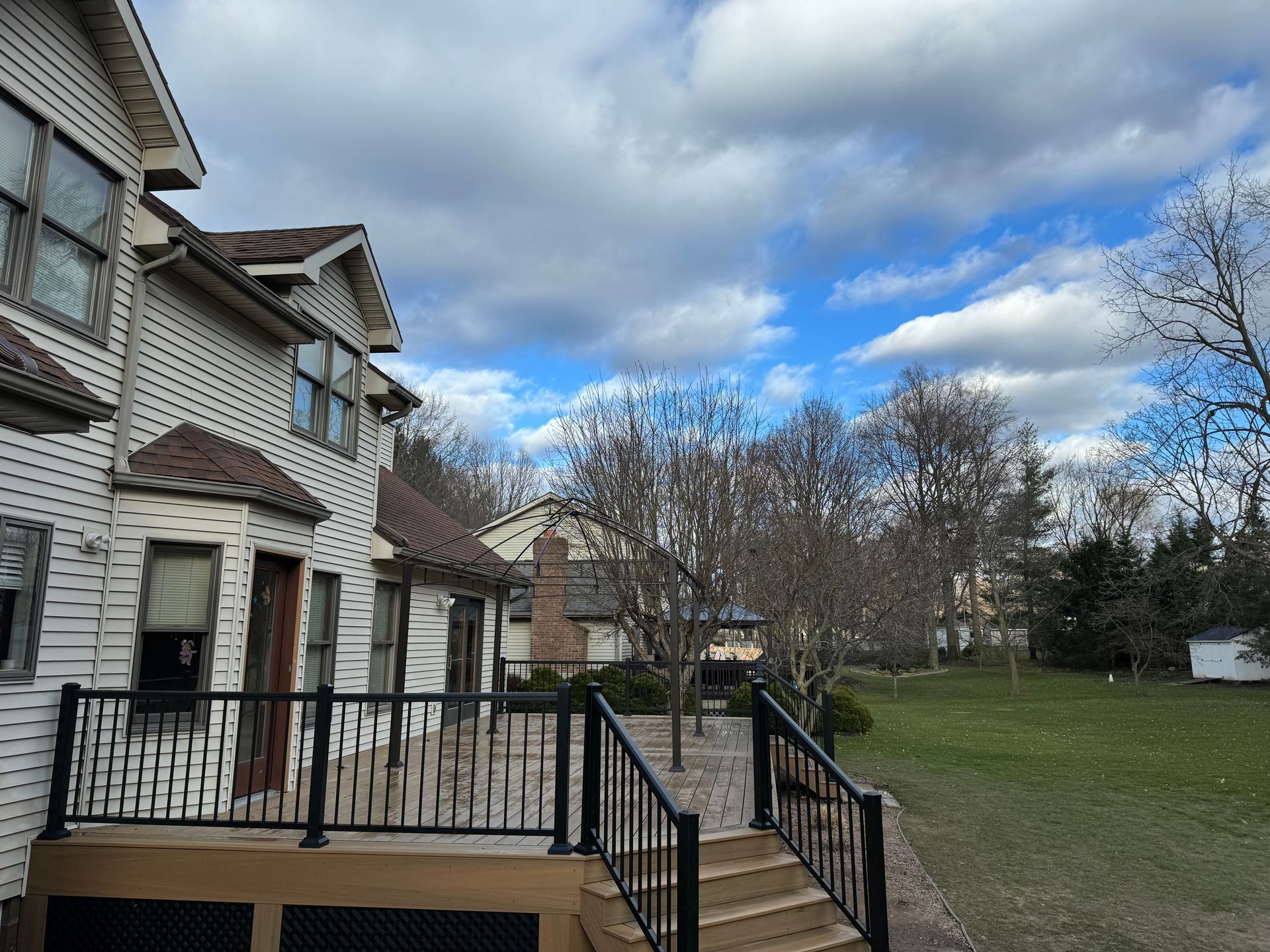 Two-story house with a deck, black railings, and a grassy yard under a cloudy blue sky.