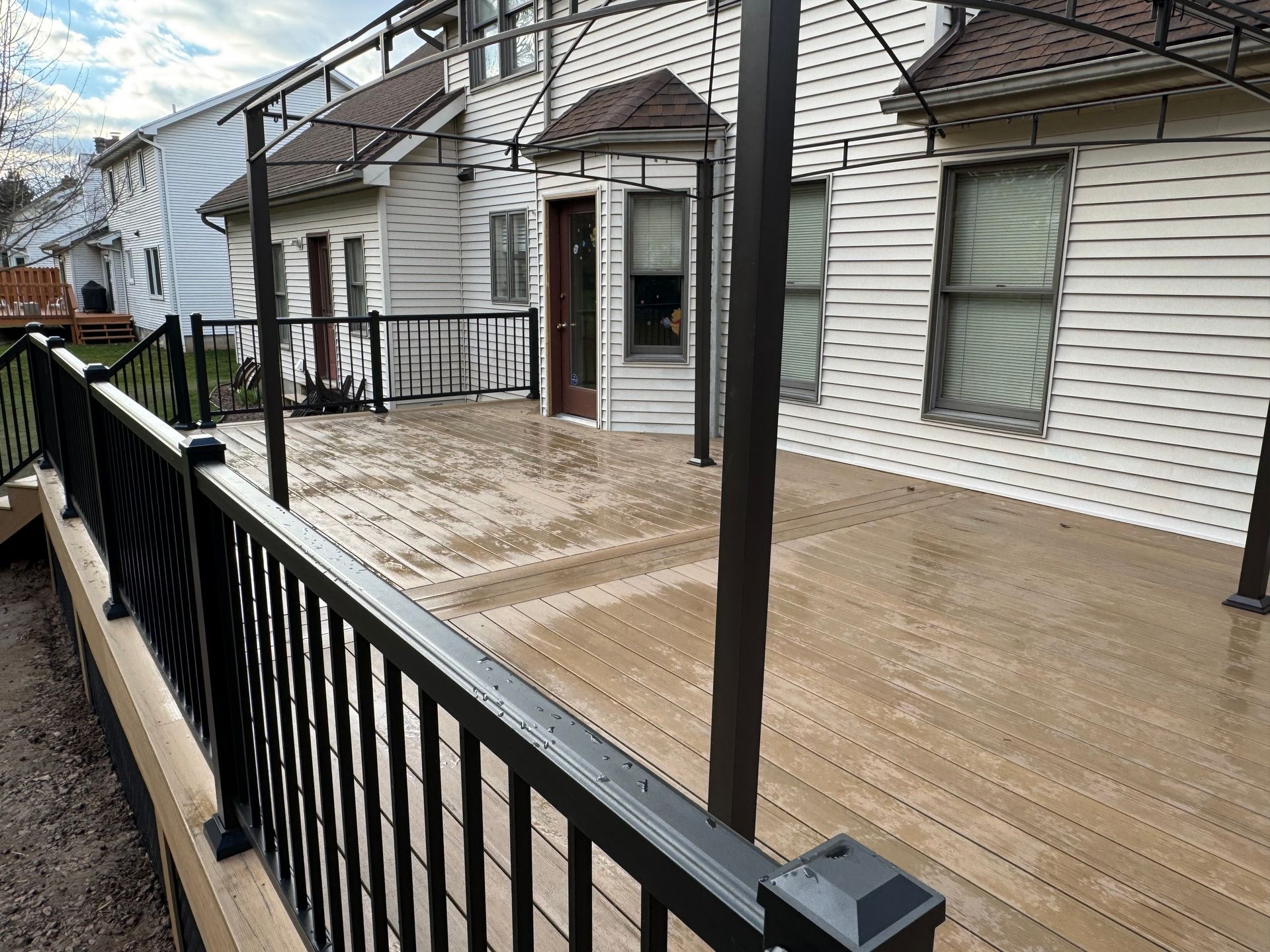 Black railing surrounds a wooden deck, with houses in the background and support columns extending upwards.