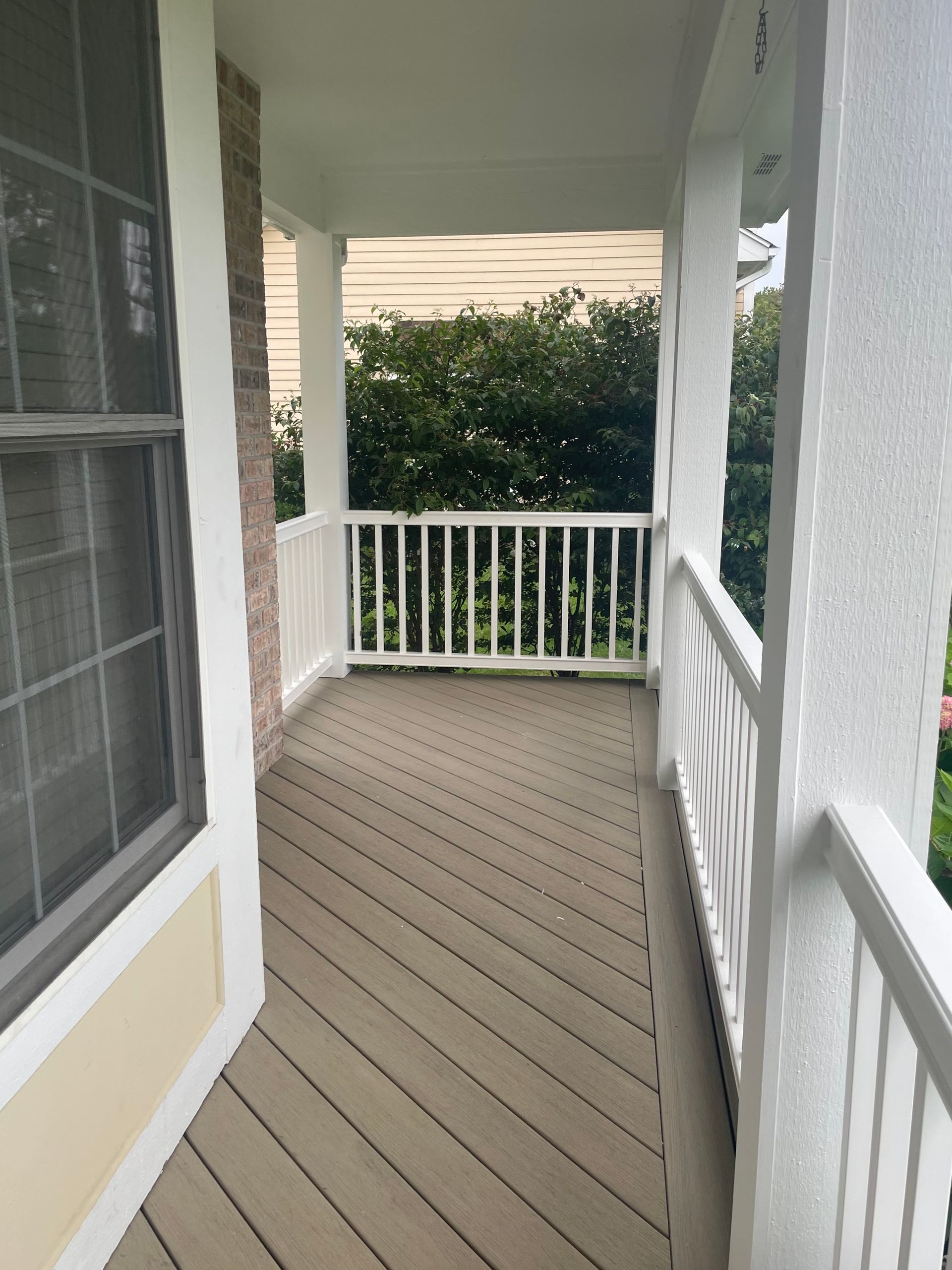 Covered porch with a wooden floor and white railing, overlooking greenery.