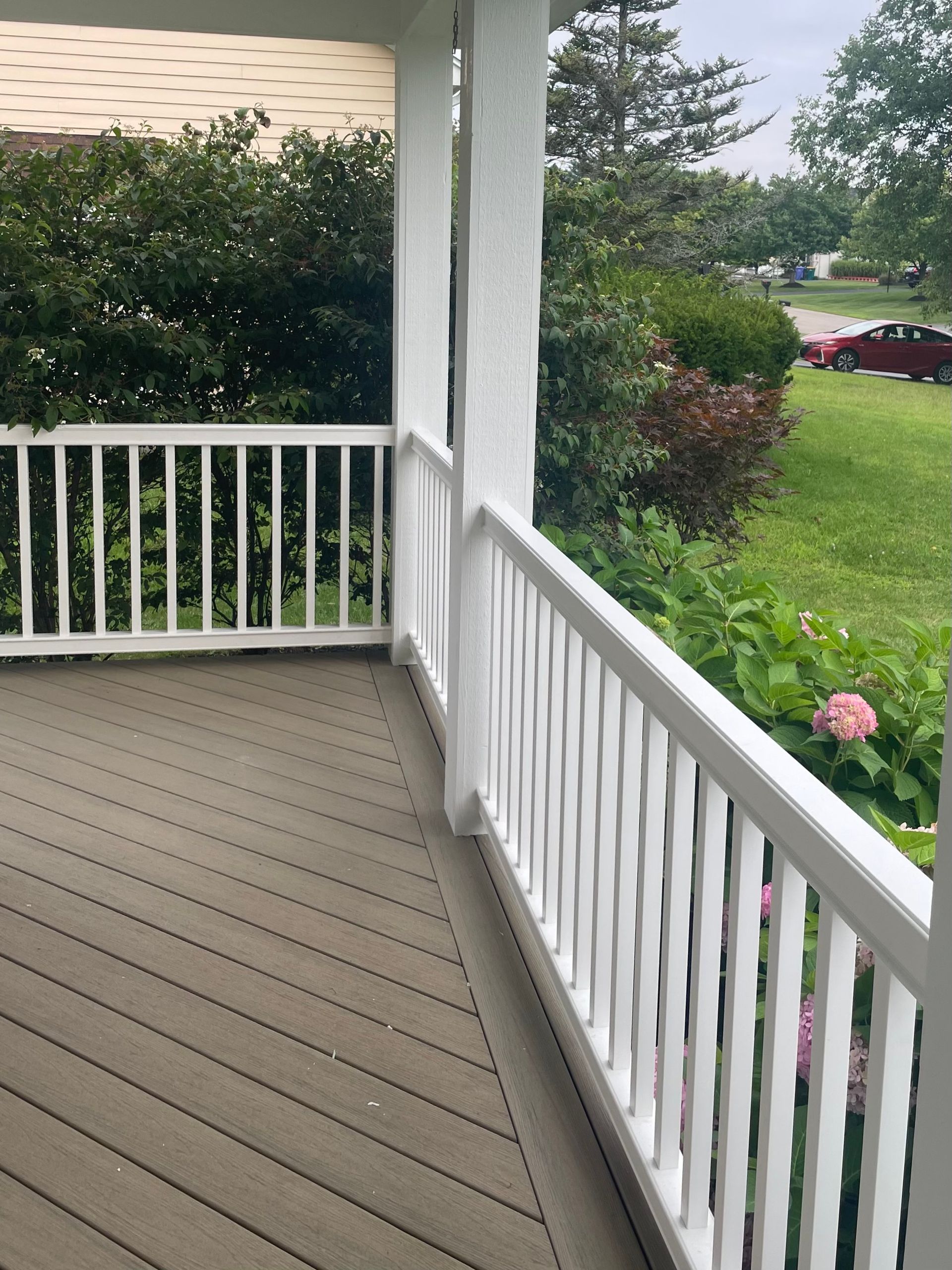 White porch railing with gray floorboards, overlooking a green yard with bushes and a car.