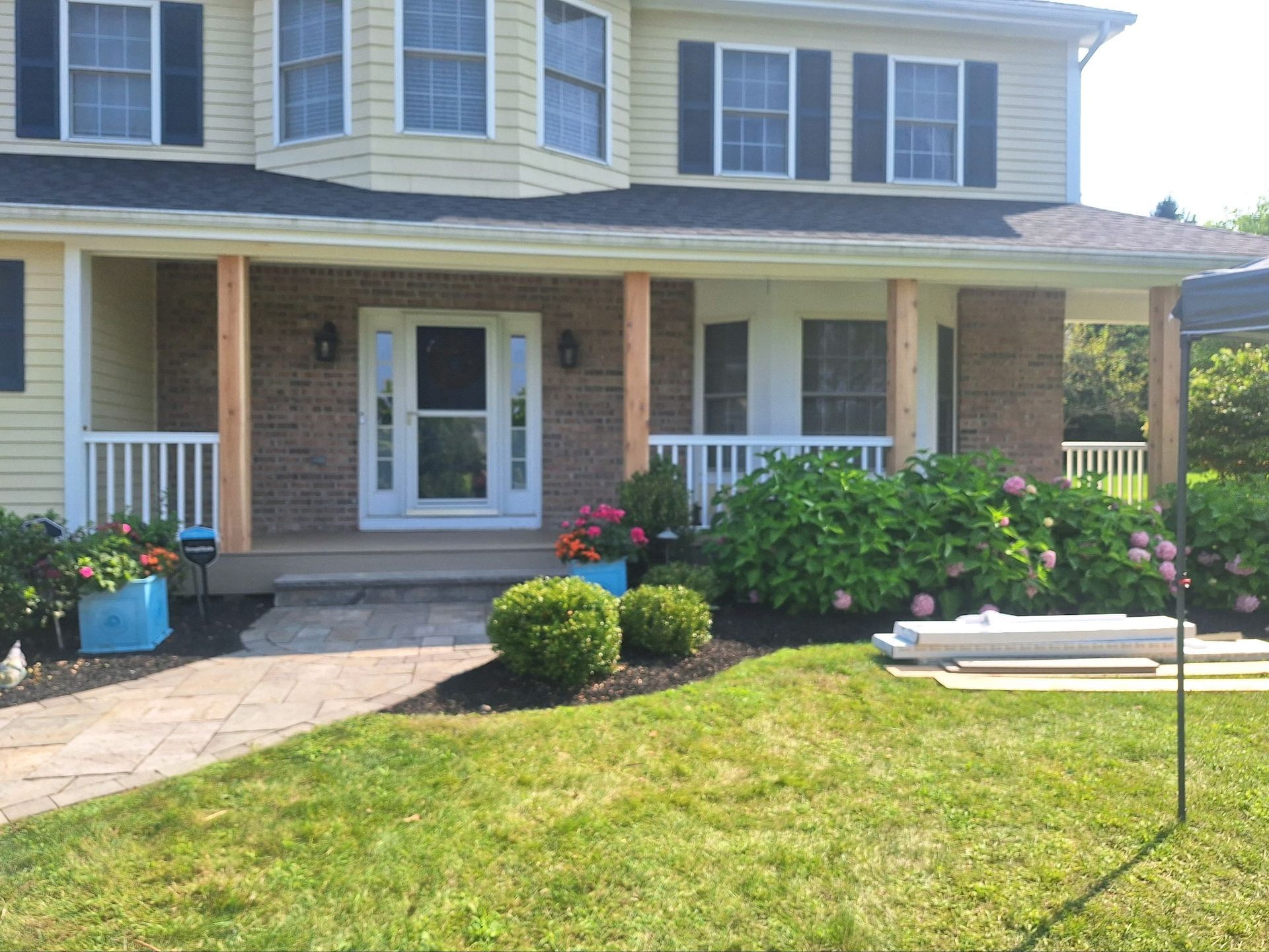 House with a front porch, featuring white railing, brick pillars, and blue flower pots, on a sunny day.