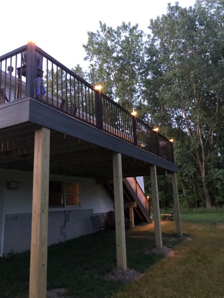 Deck with black railing, brown supports, and string lights in a wooded backyard setting.
