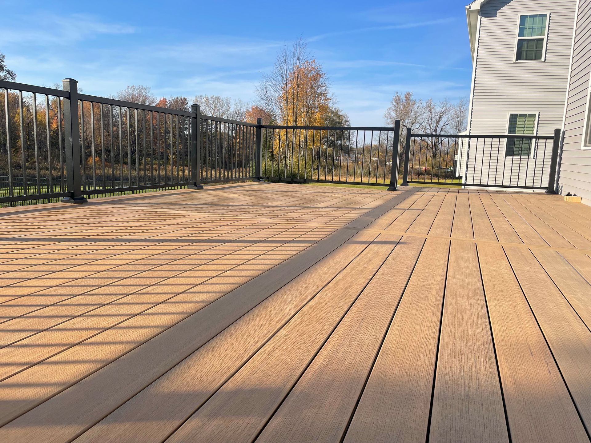 Wooden deck with black railing, against a two-story house on a sunny day.