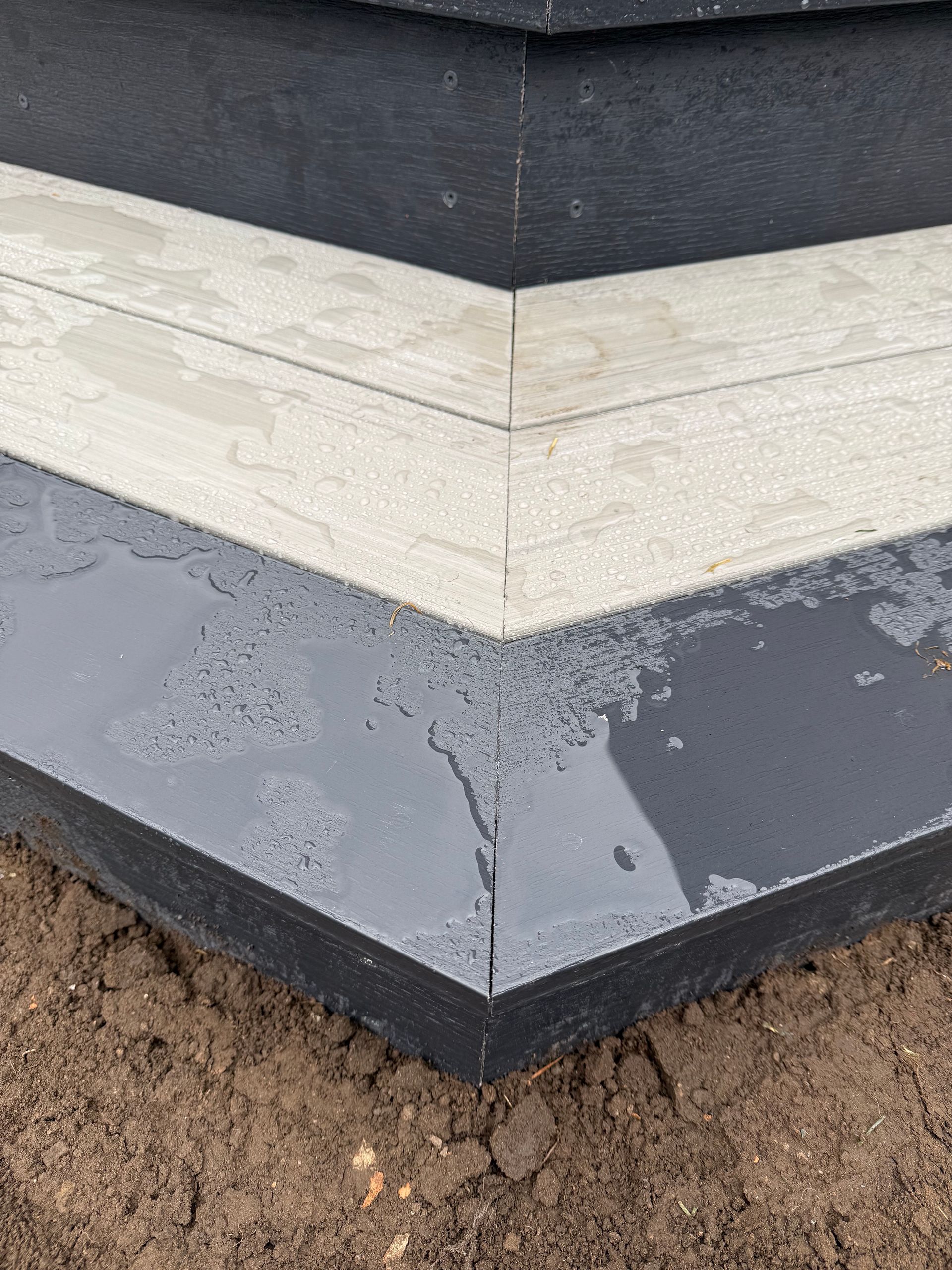Close-up of a house corner with black and white trim above a dark gray foundation over soil.
