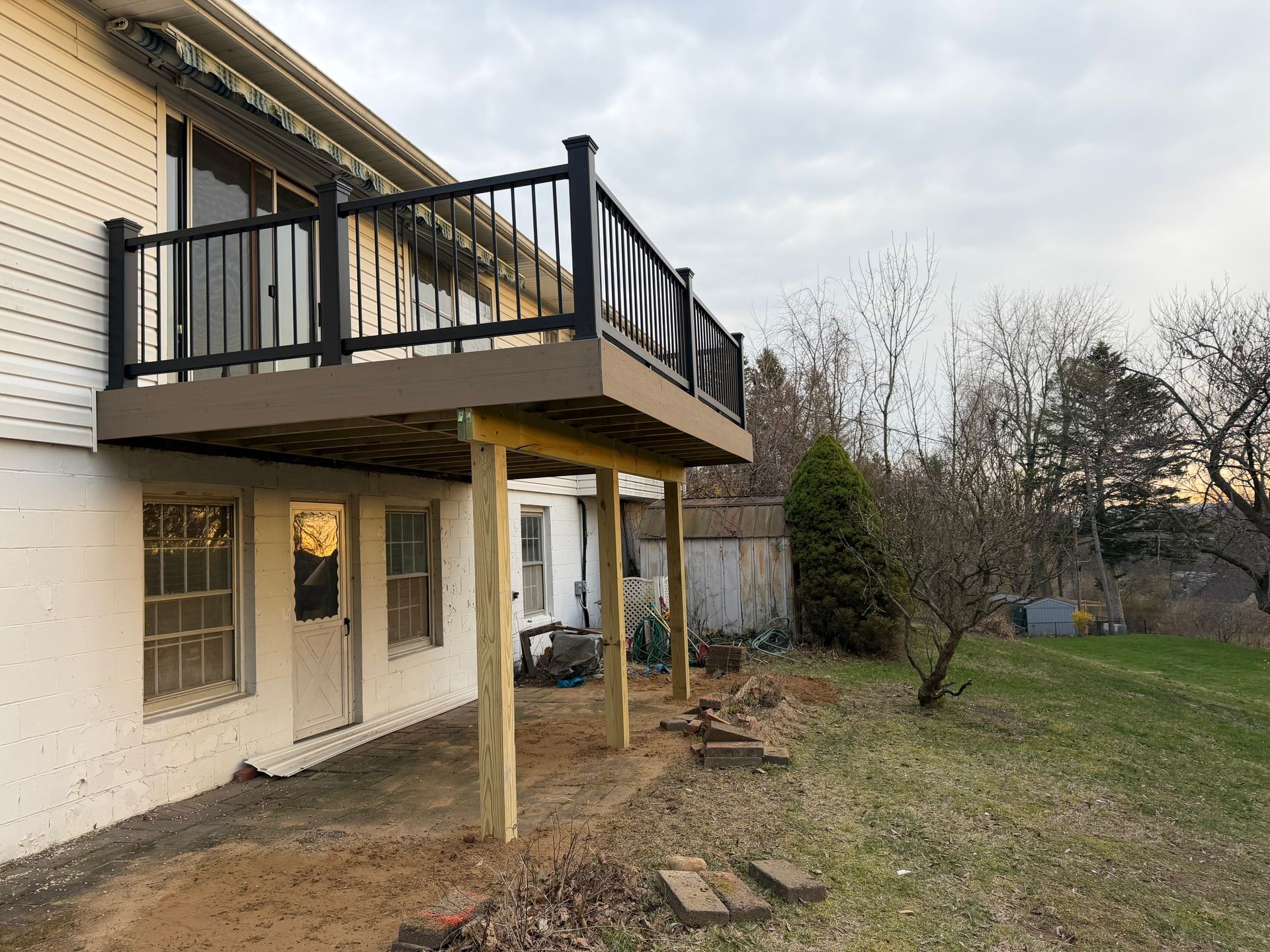 Two-story house with a raised black balcony and a dirt yard on an overcast day
