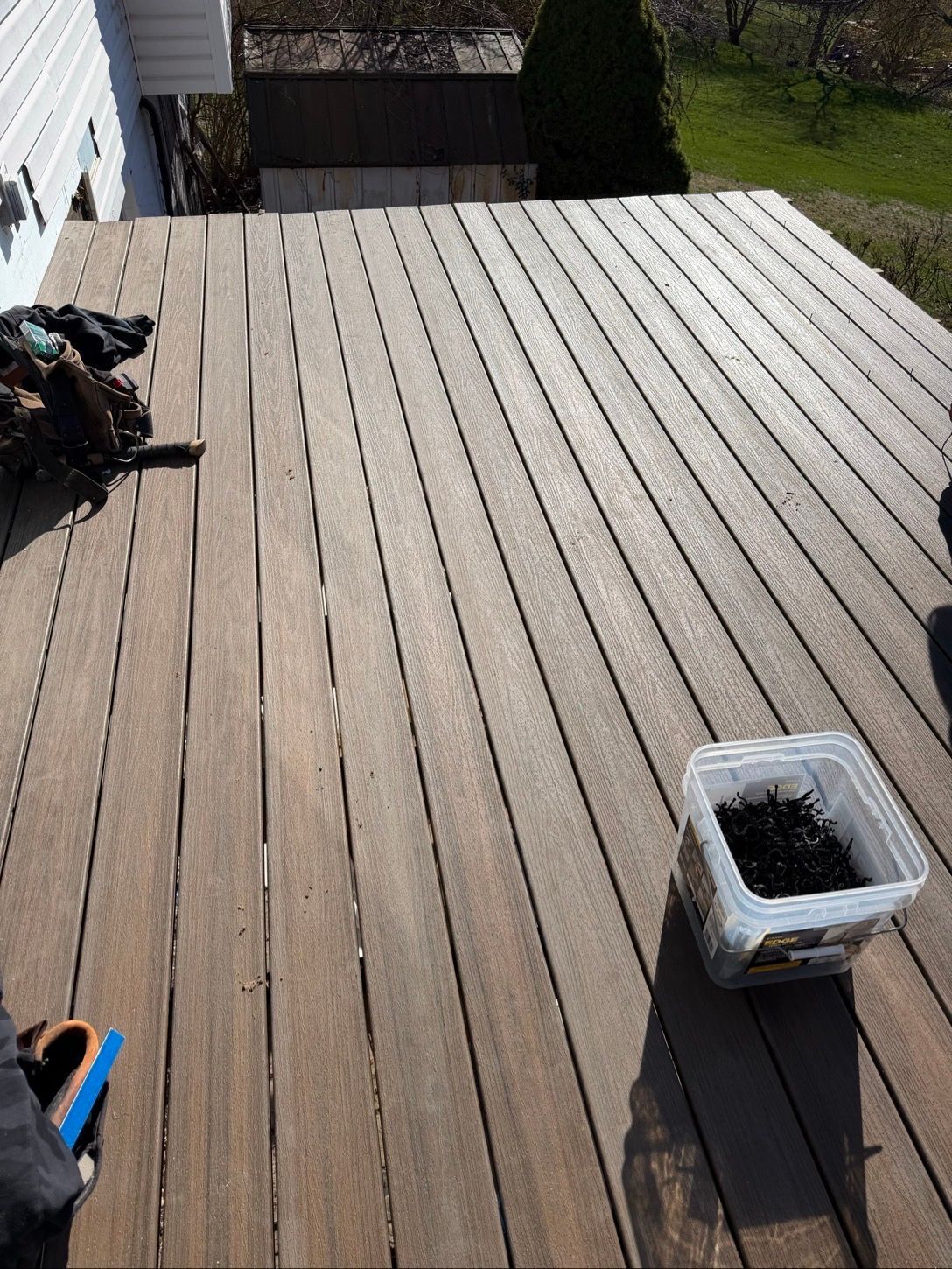 Wooden backyard deck with a planter, small tools, and a dark railing beside a grassy yard