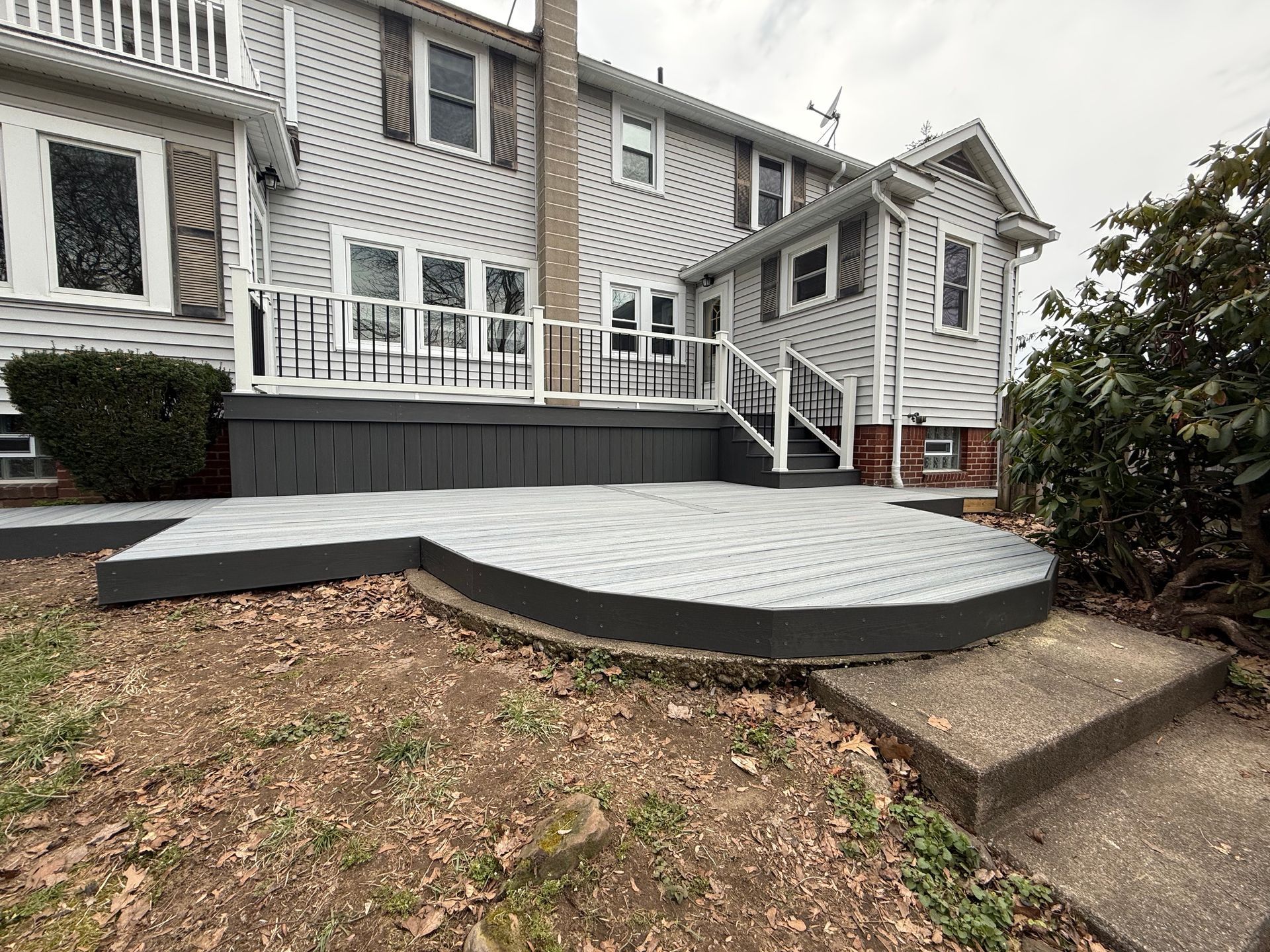 Raised gray brick patio with railing and white stairs beside a house, bordered by mulch and bushes