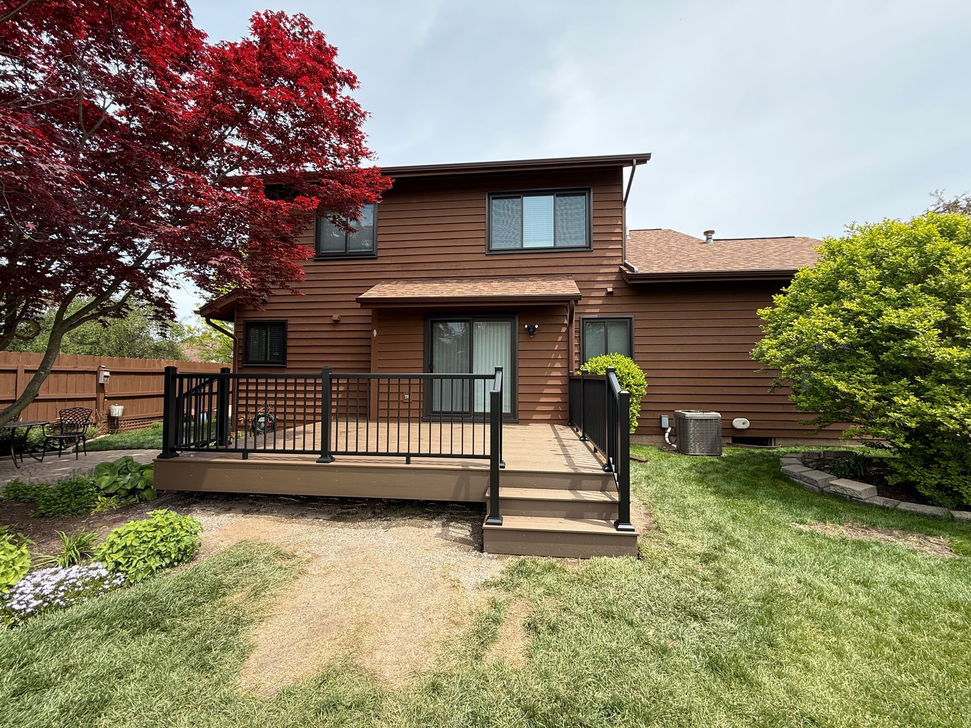 Two-story brown house with a front deck, black railing, and red tree in a grassy yard