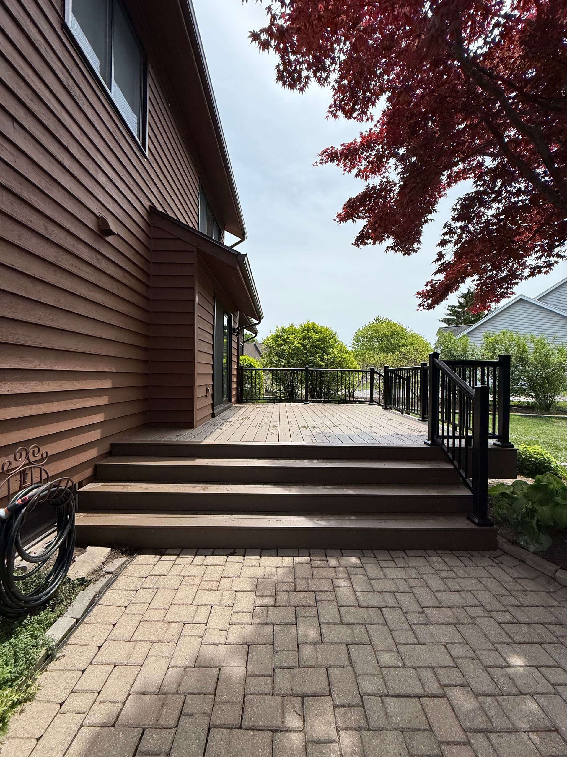 Brick patio steps beside a brown-sided house, leading to a fenced terrace with trees beyond.