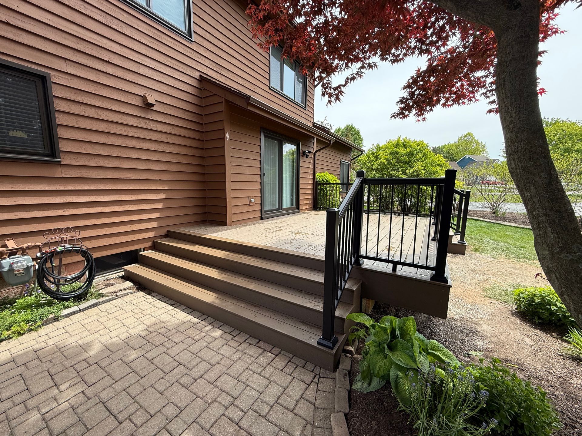 Brick house entrance with steps, black railing, and a paver walkway beside a tree and garden bed