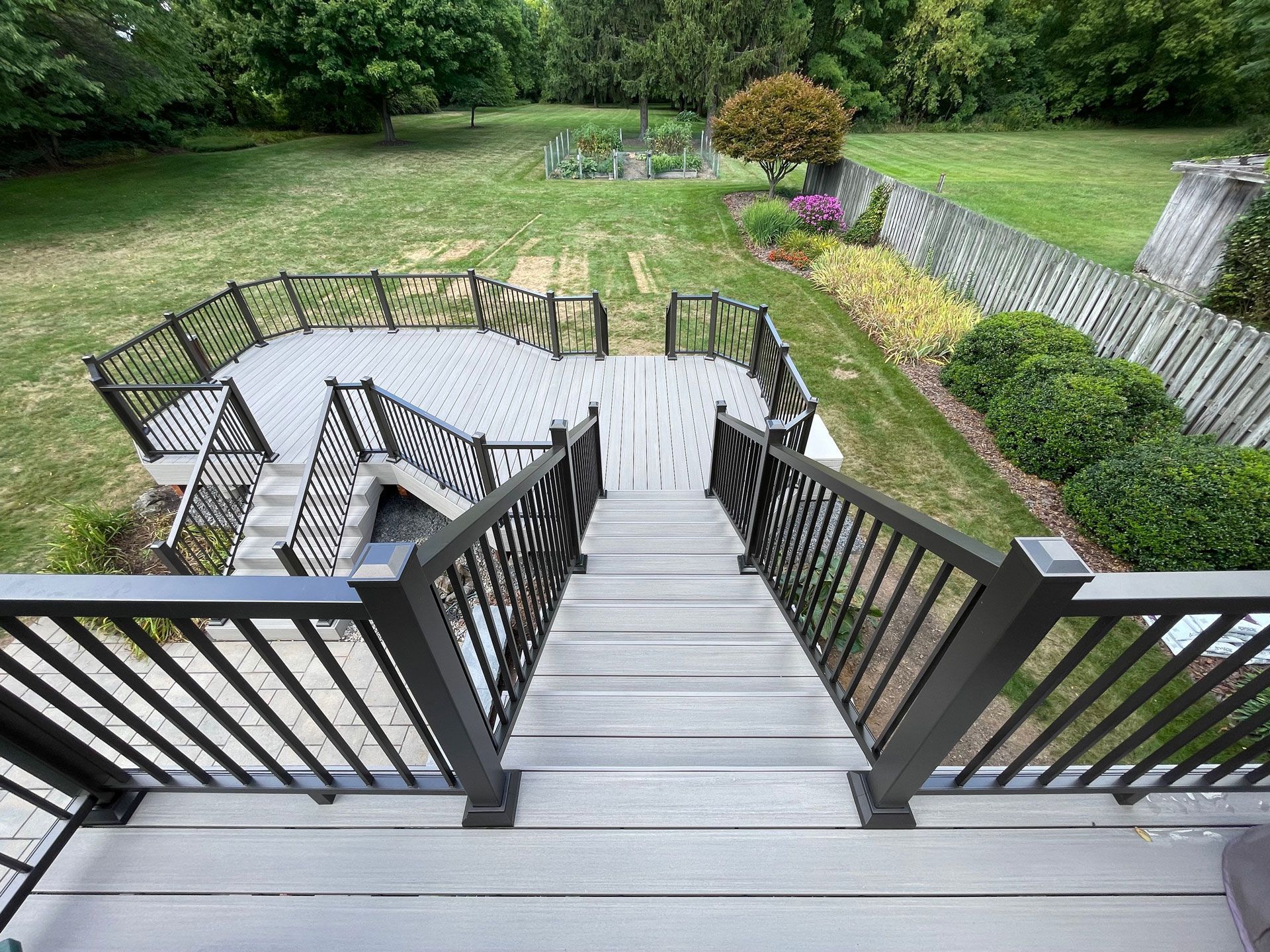 Grey deck with decorative railings, steps lead to backyard with garden and trees.