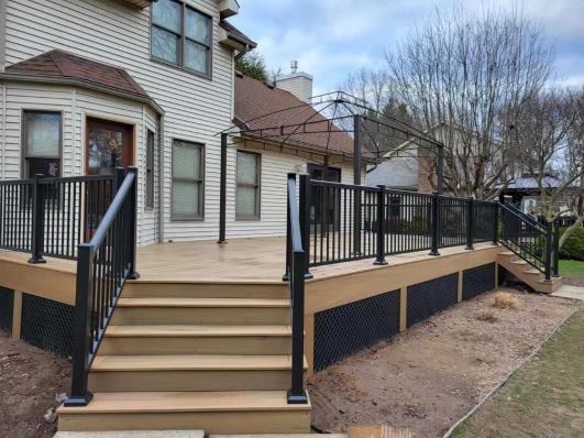 Black railing surrounds a multi-level deck with stairs leading to a light brown deck. House in background.