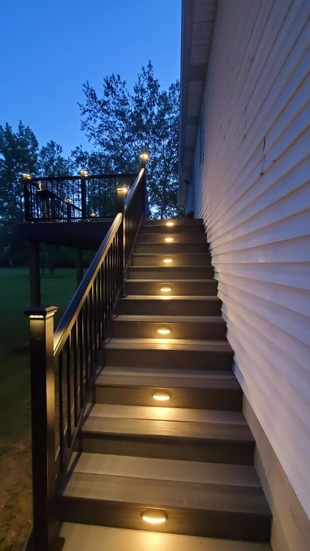 Exterior stairs with embedded lights leading to a dark deck, against a white house siding, at dusk.