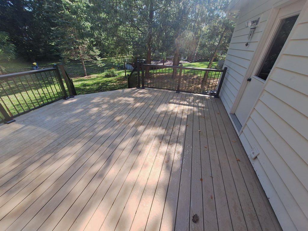 Wooden deck with black railing, attached to a white house with a door, overlooking a grassy area and trees.