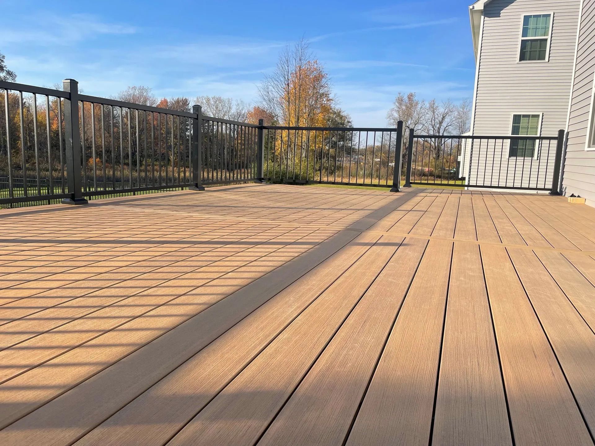 Wooden deck with black metal railing, autumn trees, and a house in the background under a clear blue sky