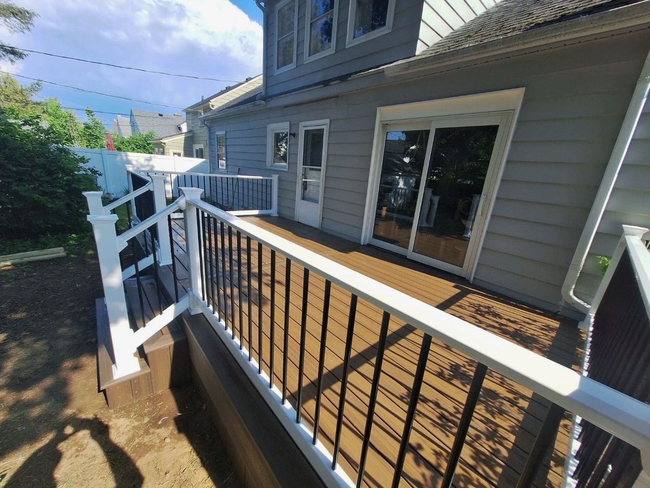 Backyard deck with brown composite decking, white railing, and black balusters. House with sliding glass door in background.