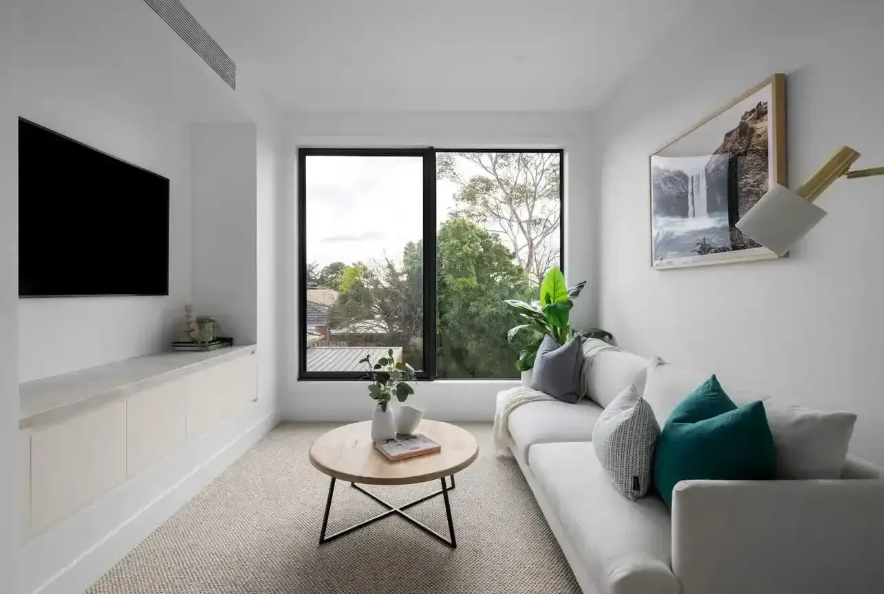 Living room with white walls, white sofa, black-framed window overlooking trees, and TV on wall.
