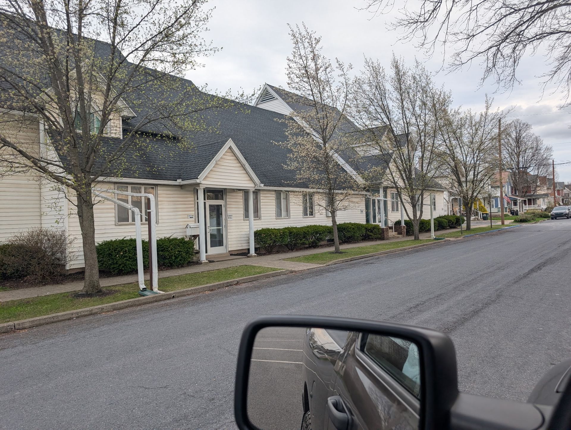 Quiet residential street with beige houses, bare trees, and a car side mirror in the foreground