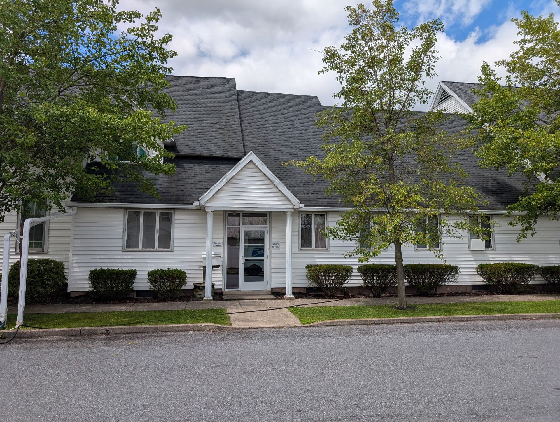 White suburban building with gray roof, front entrance, and trees along a street under a cloudy sky