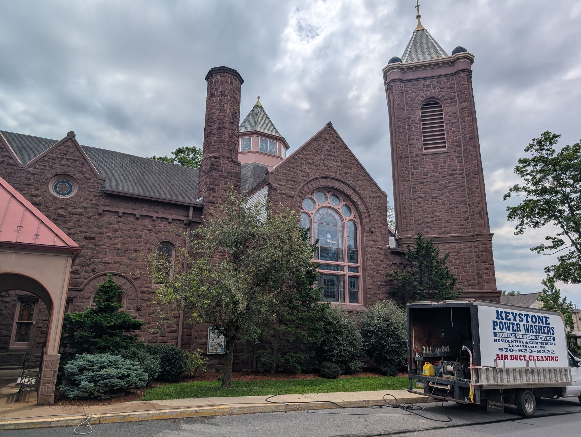 Brick church with arched stained-glass window and tower under cloudy sky, van parked outside