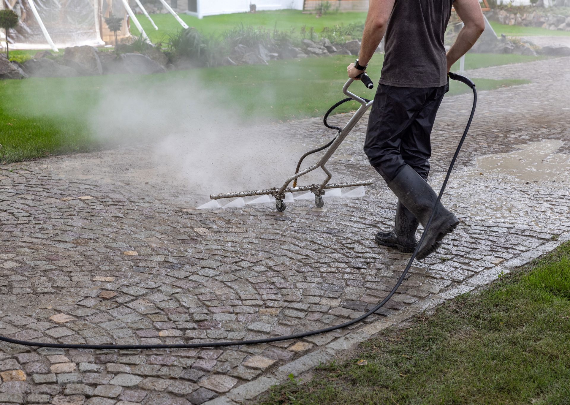 Person power-washing a cobblestone patio beside a lawn and hedge