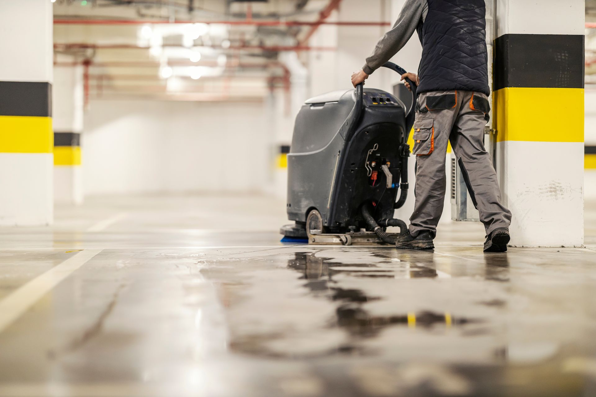 Person cleaning a wet parking garage floor with a floor scrubber beside yellow-black columns