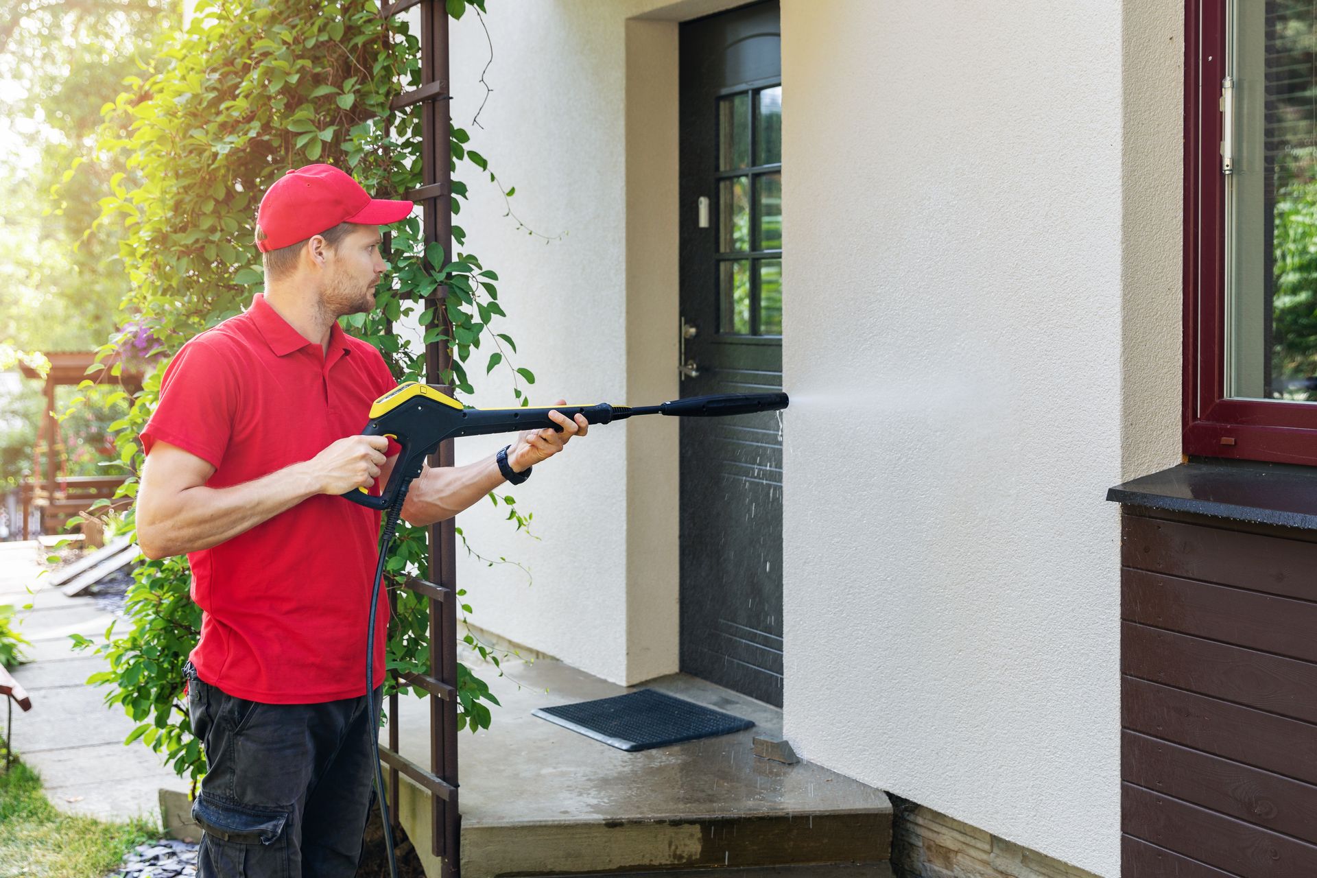 Person in red shirt and cap pressure-washing a house wall beside a doorway and window