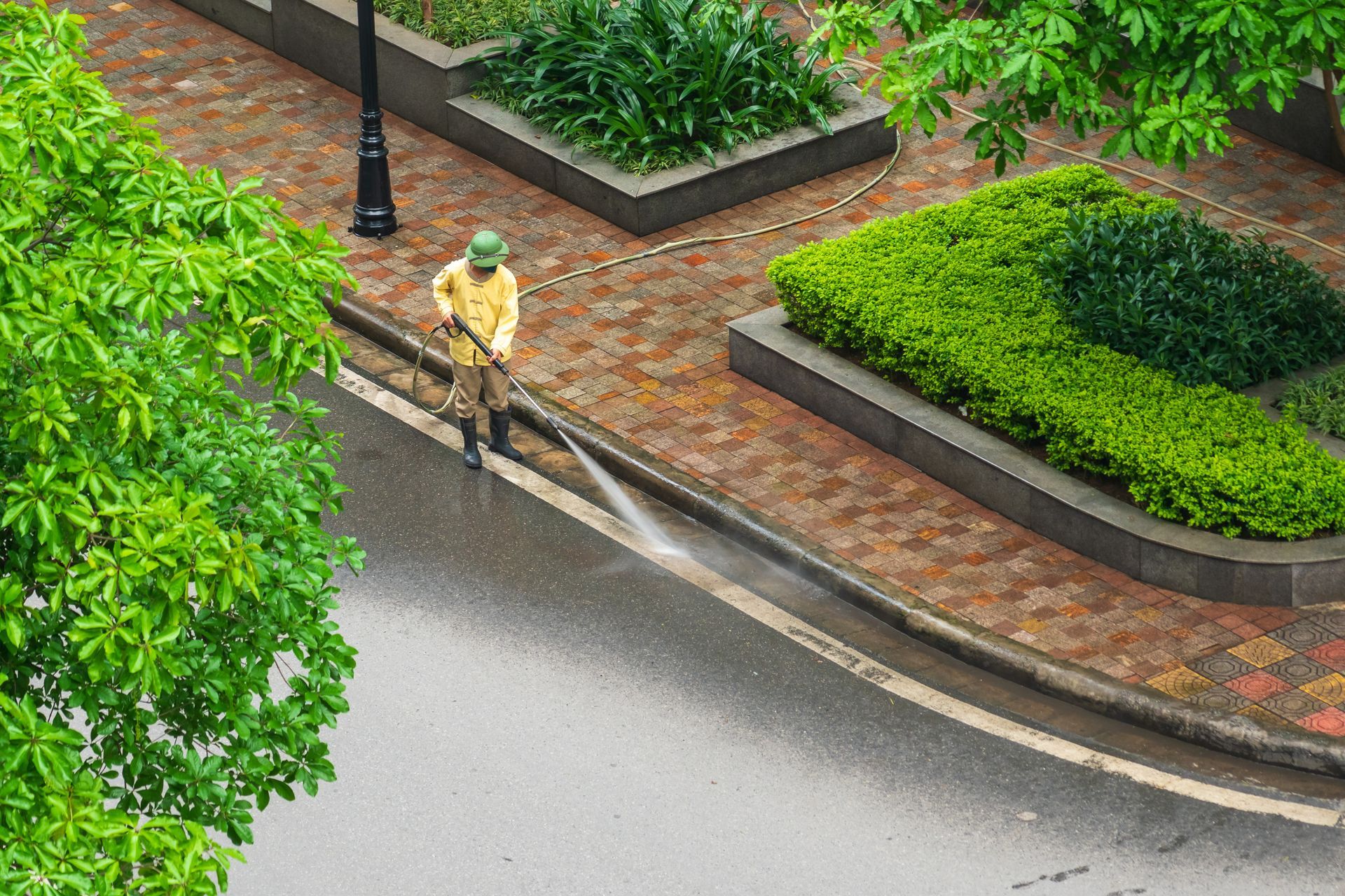 Worker using a leaf blower on a wet street beside a brick sidewalk and green hedges