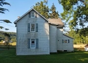 White farmhouse with blue shutters and attached addition, standing on a grassy yard under trees.