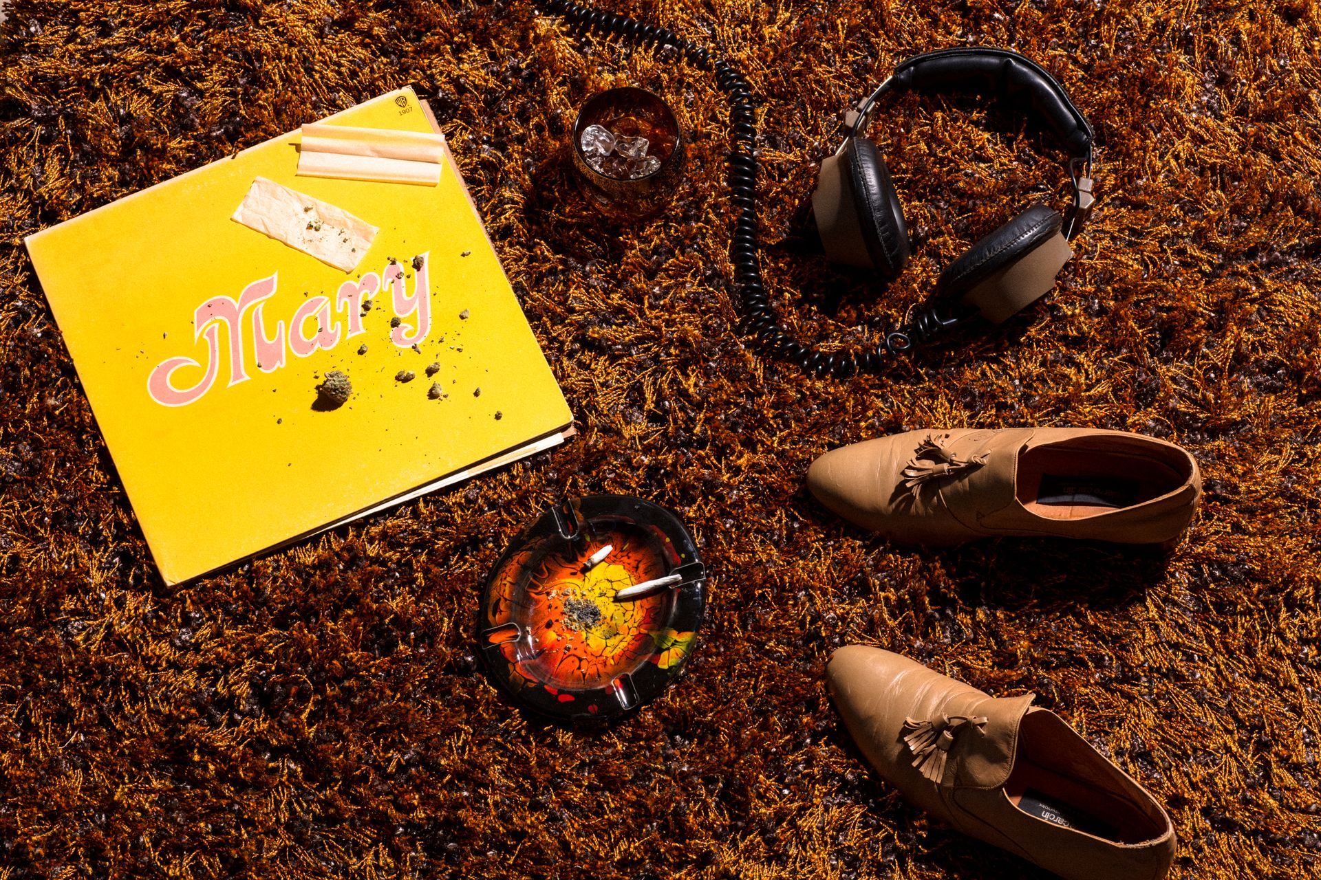 Yellow book, sunglasses, wristwatch, and tan shoes arranged on a brown textured surface.