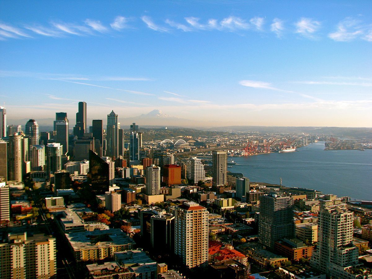 City skyline with tall buildings, waterfront, blue sky with wispy clouds, and distant mountains.
