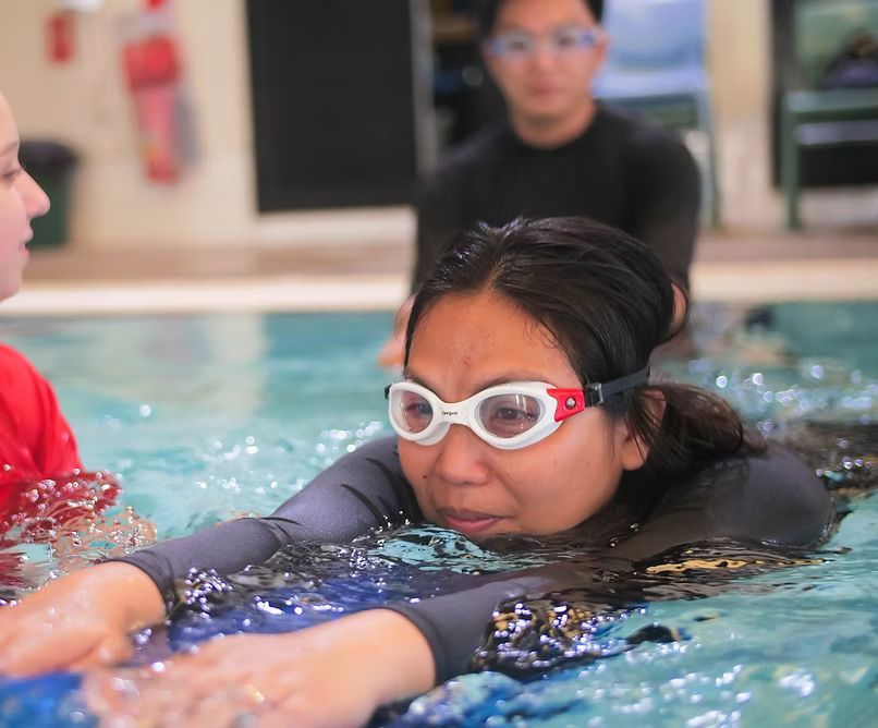 Woman in goggles swims in a pool, holding a blue board, with a person behind her.