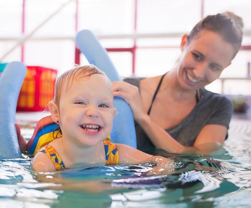 Child in pool with flotation device smiles at instructor in a pool setting.