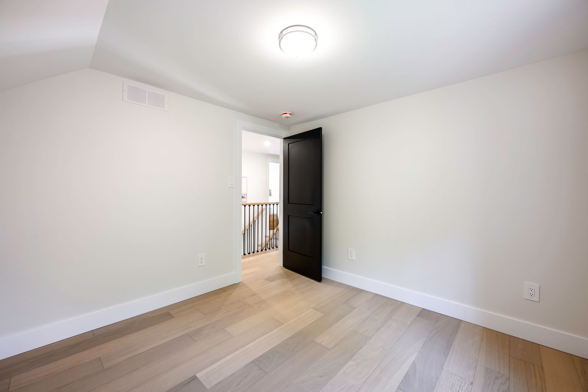Empty room with light wood floor, white walls, black door, and a ceiling light.