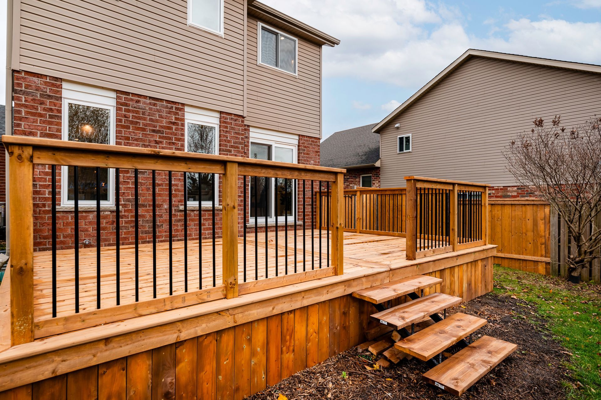Wooden deck with stairs, black railing, and connecting to a two-story brick and beige house.