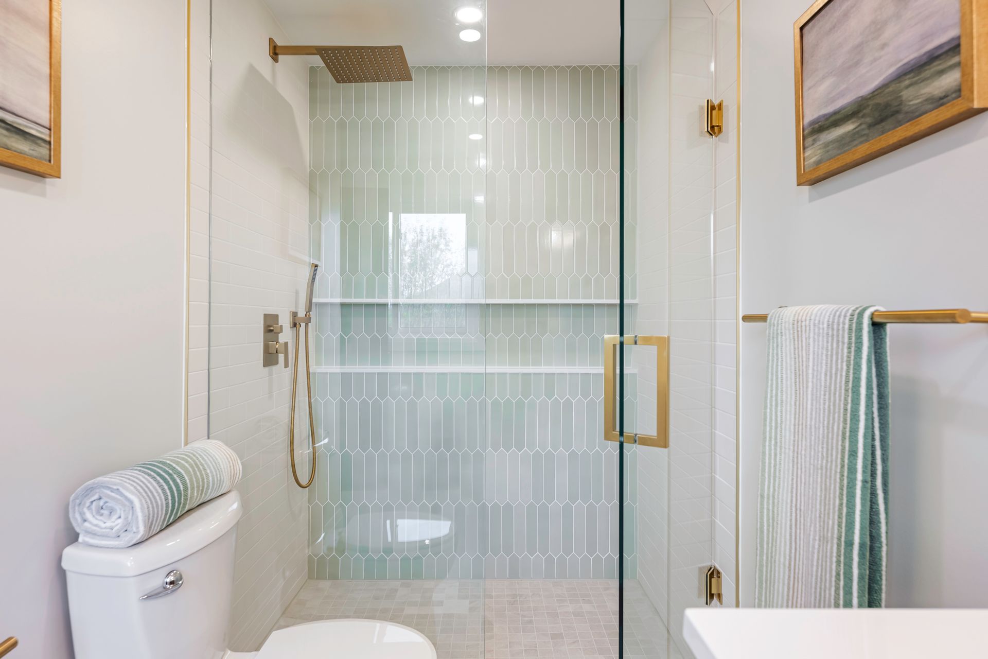 Modern bathroom with glass shower, gold fixtures, and pale green tile. White walls and toilet.