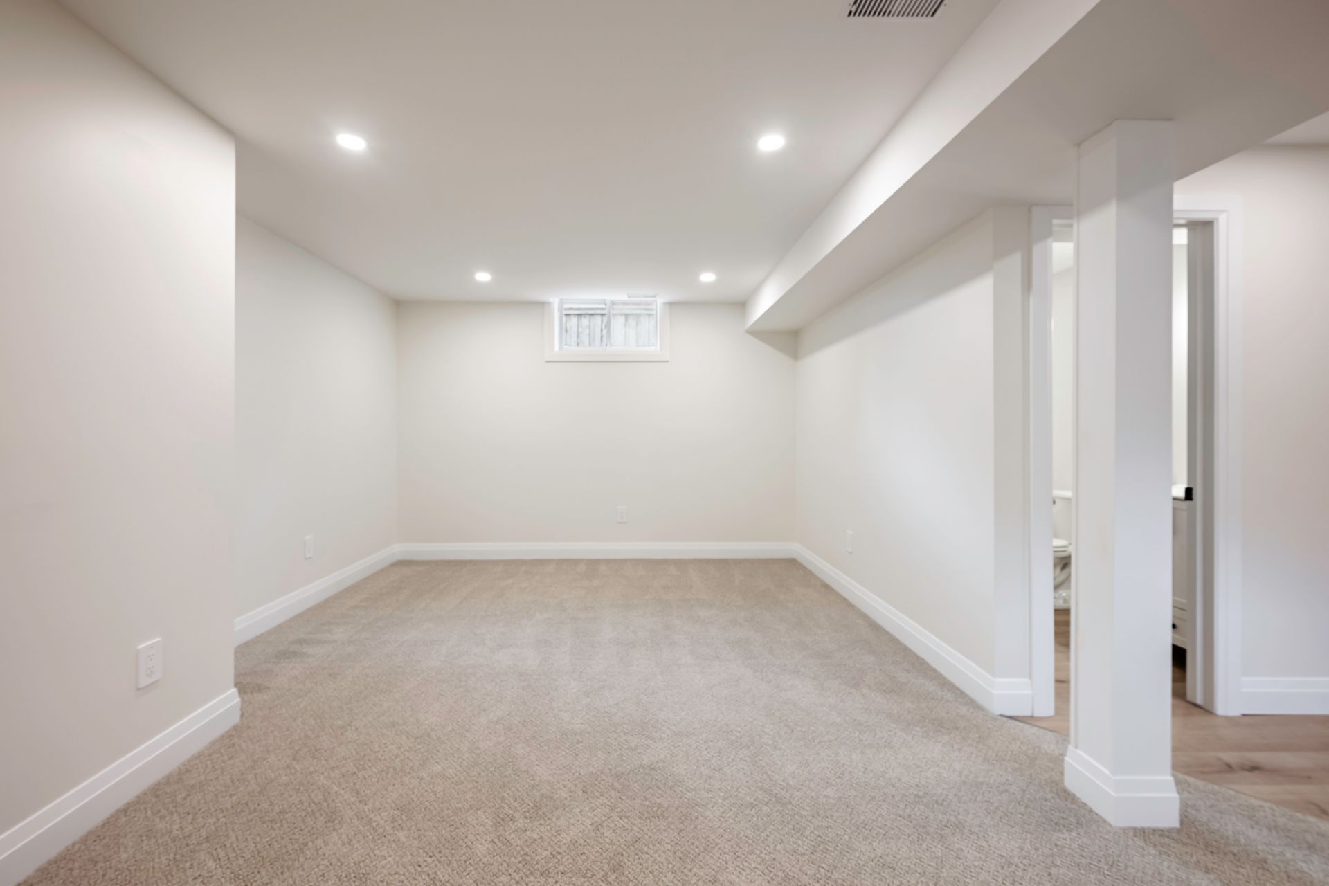 Empty, finished basement room with beige carpet, white walls, and recessed lighting. A doorway leads to another room.