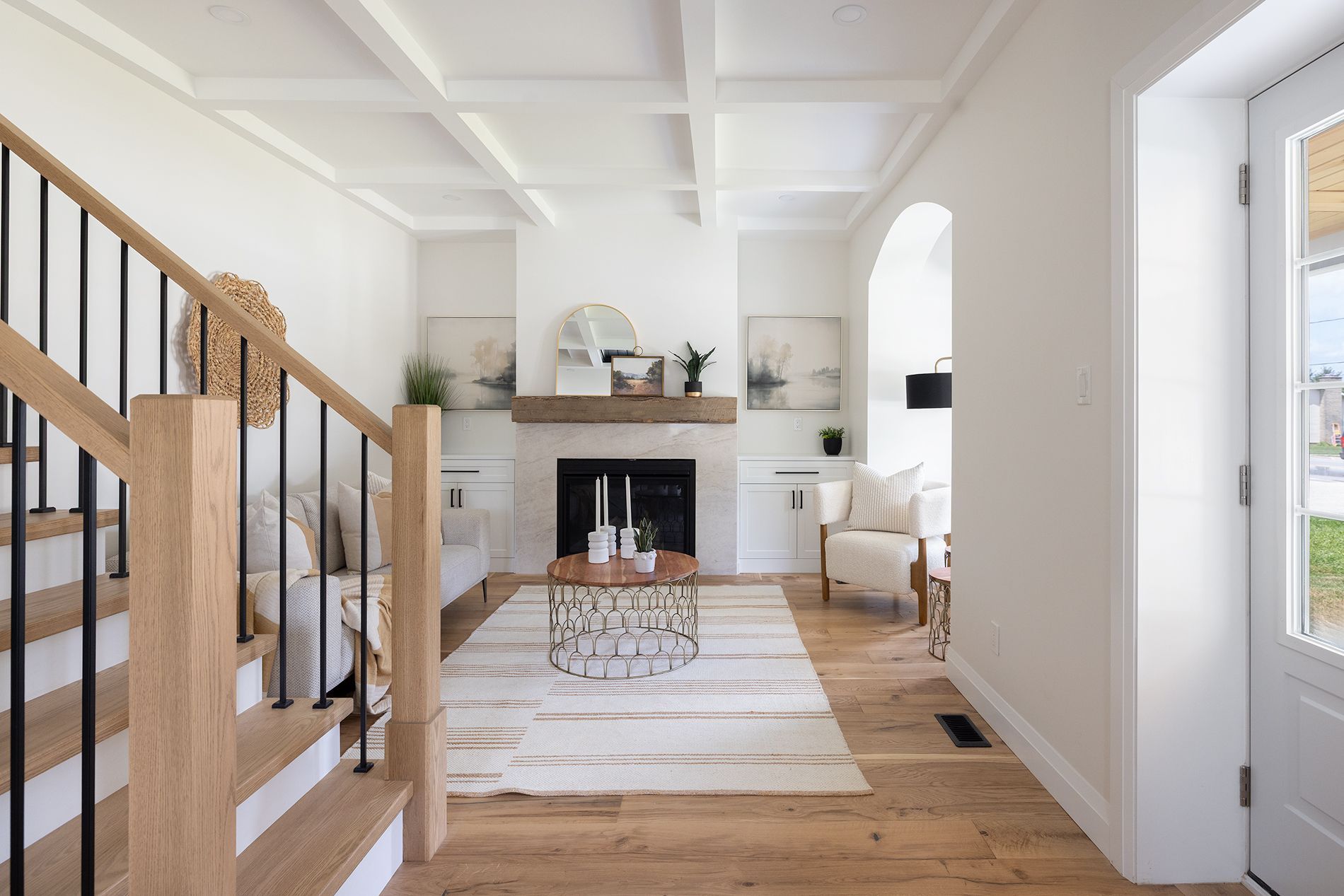 Bright living room with fireplace, wood floors, staircase, and white walls.